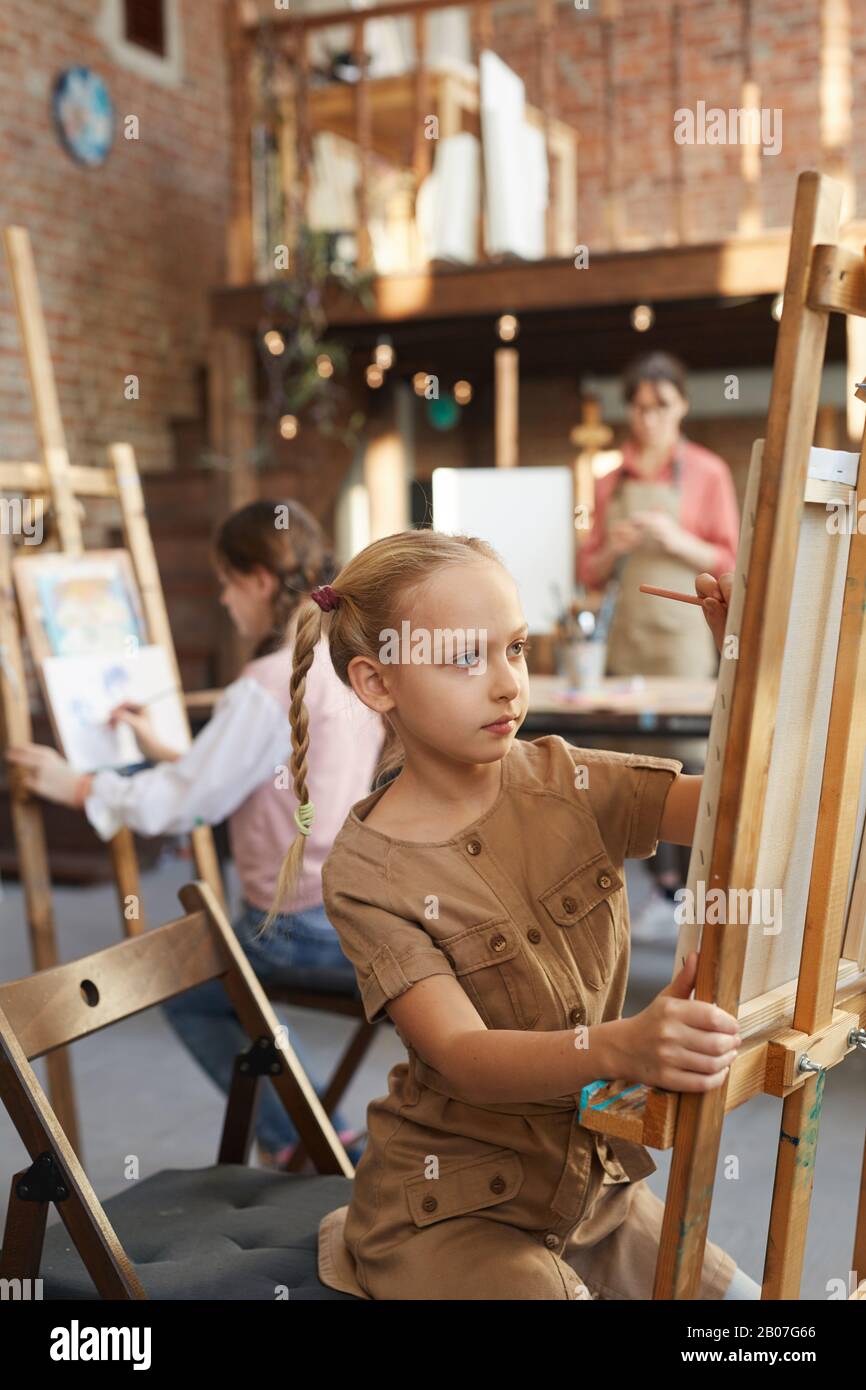 Kleines Mädchen, das während des Kunstunterrichts im Kunststudio vor der Staffelei sitzt und mit anderen Studenten malt Stockfoto