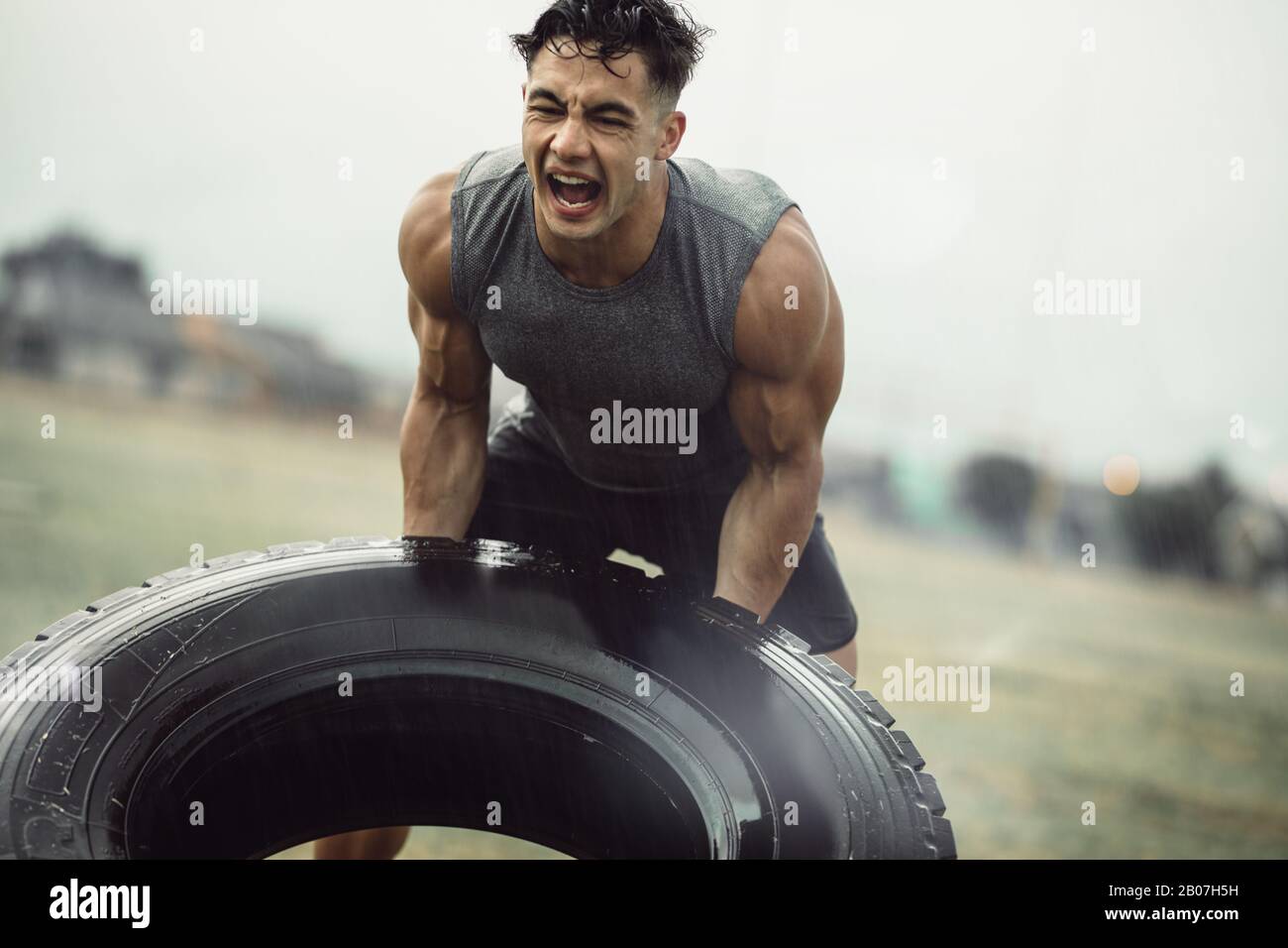 Harter junger Athlet, der im Regen eine Übung zum Spiegeln des Reifens macht. Muskulöser Mann macht Cross-Training im Freien auf dem Feld. Stockfoto