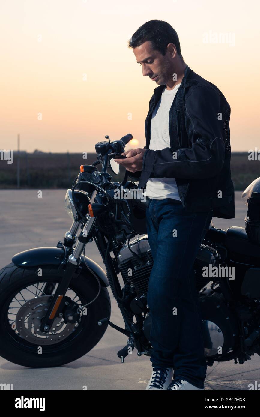 Latin man schaut bei Sonnenuntergang auf das Smartphone neben seinem eigenen Motorrad. Stockfoto