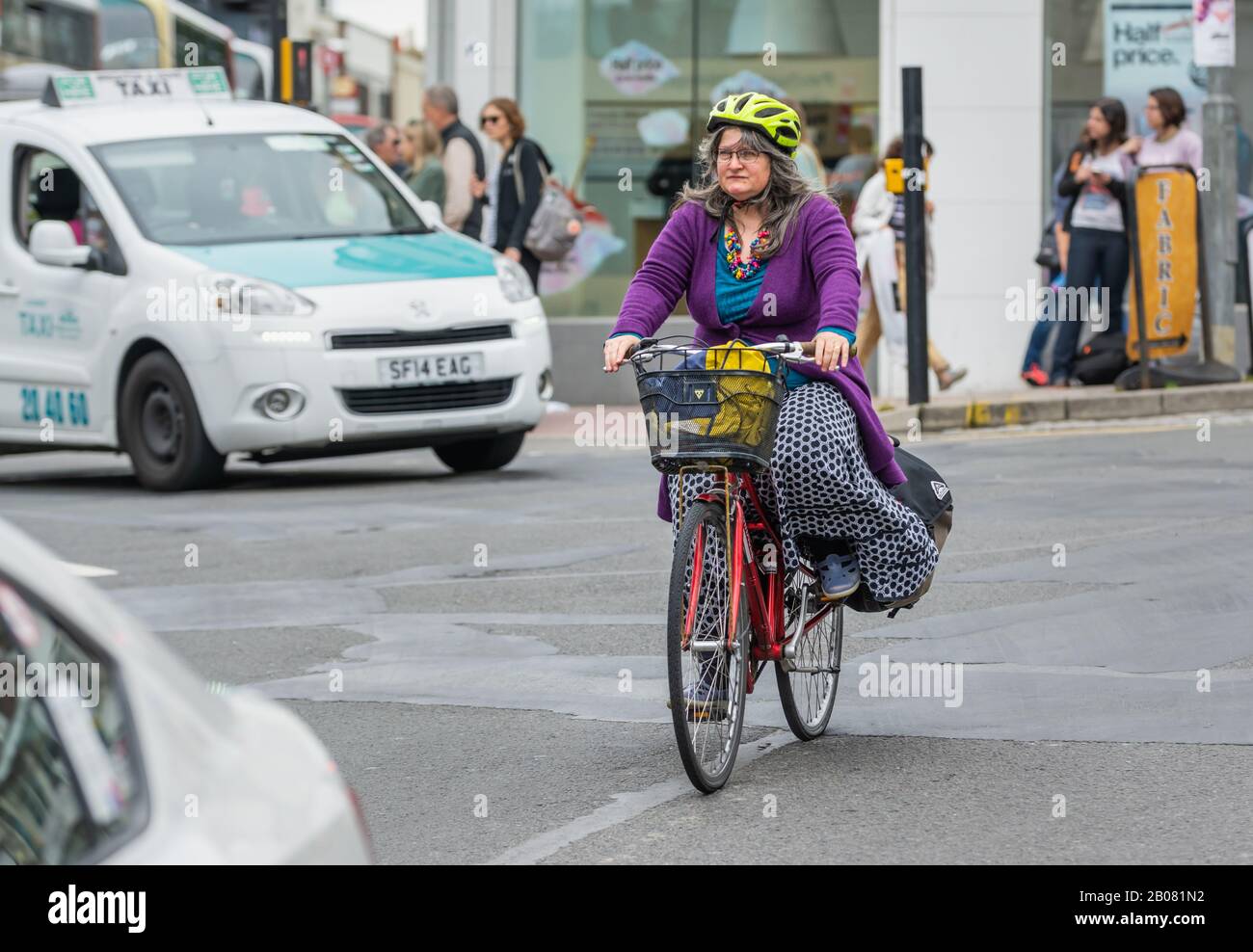 Radfahrerin mittleren Alters, die einen Helm trägt, mit dem Fahrrad auf einer Straße in der geschäftigen Stadt Brighton, England, Großbritannien. Frau, die mit dem Fahrrad unterwegs ist. Stockfoto