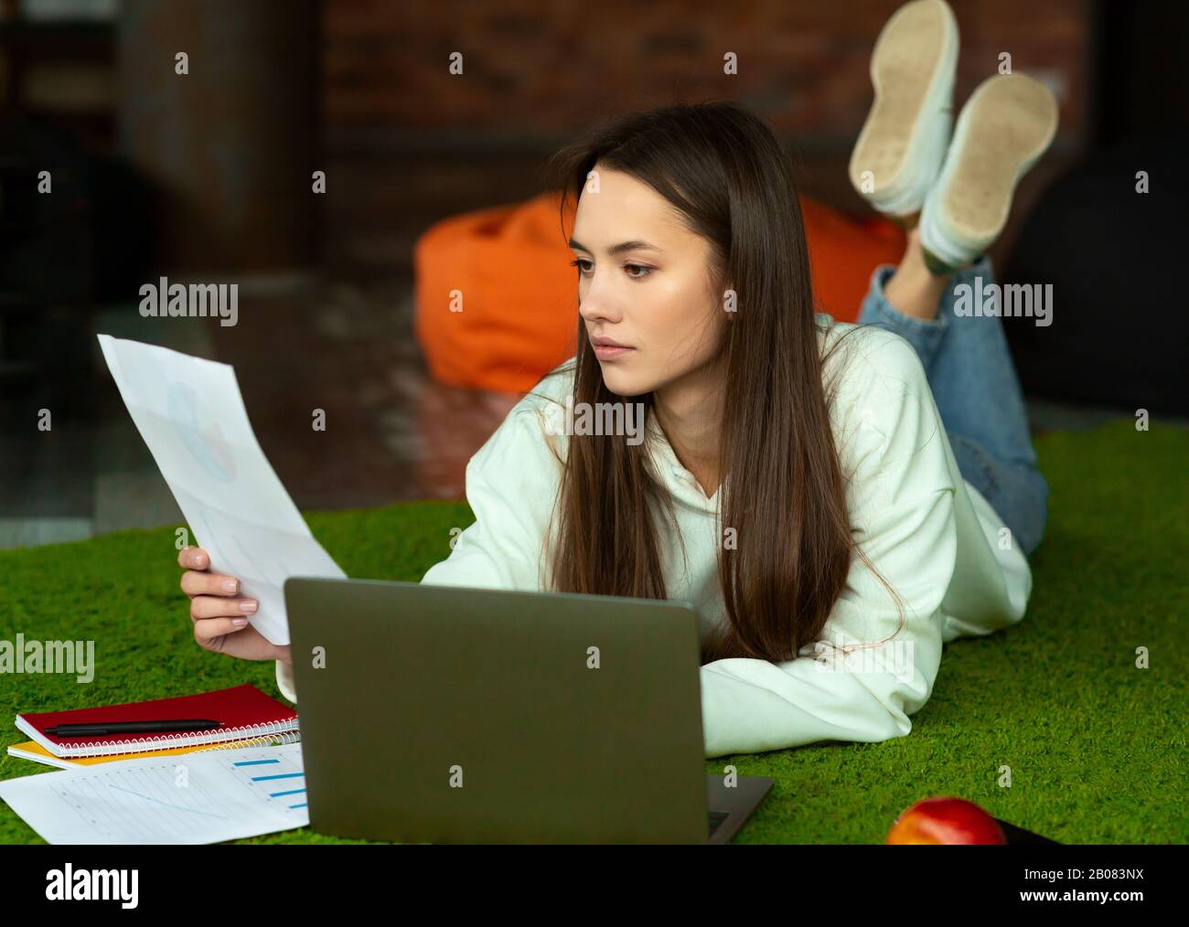 Das schöne Mädchen liegt auf dem Boden, sieht Papier an, arbeitet mit Computer und Notizblock Stockfoto