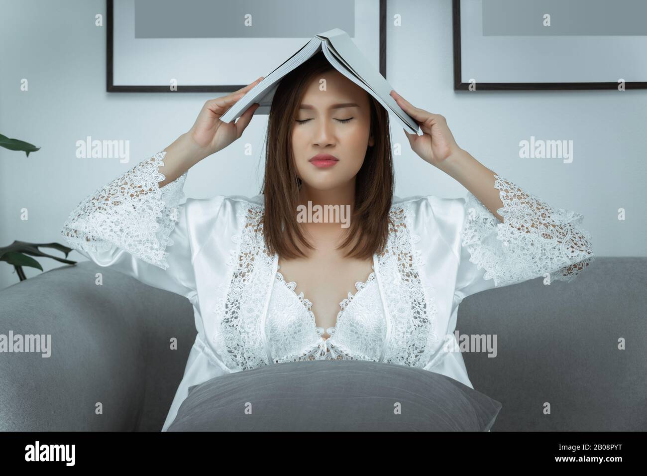 Asiatische Frauen tragen weiße Nachtkleid- und langärmlige Satin-Robe mit Blumenspitze, die Augenbelastung leidet Lesebuch sitzt auf einer Couch in der Nacht zu Hause. Stockfoto