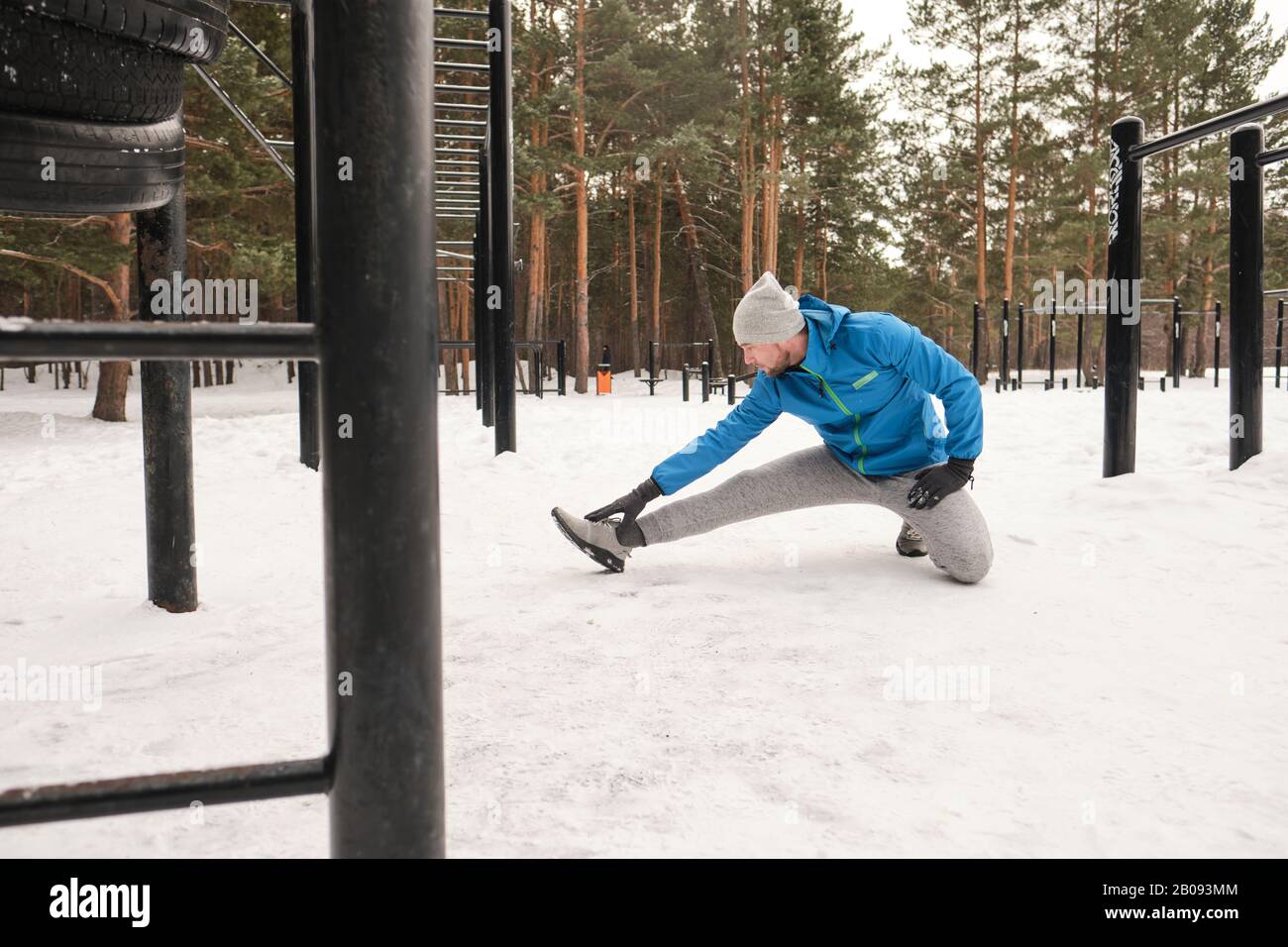 Junger Mann in blauer Jacke, der auf einem Knie sitzt und das Bein in der Kräuselposition streckt, während er im Winter-Trainingsbereich trainiert Stockfoto