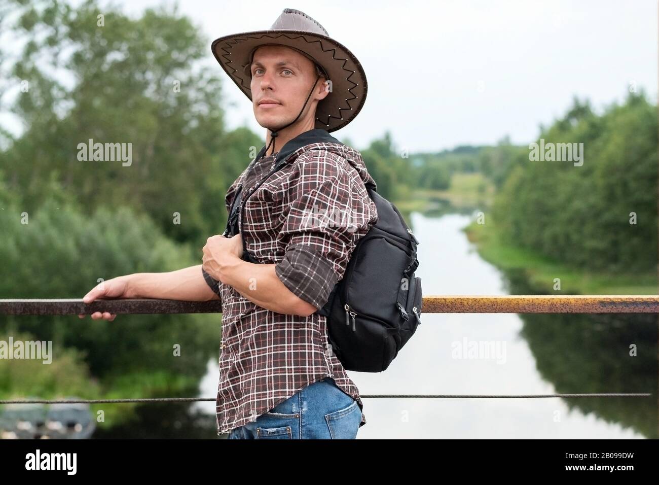 Ein Mann in einem Cowboyhut, mit einem Rucksack auf der Brücke über den Fluss. Stockfoto