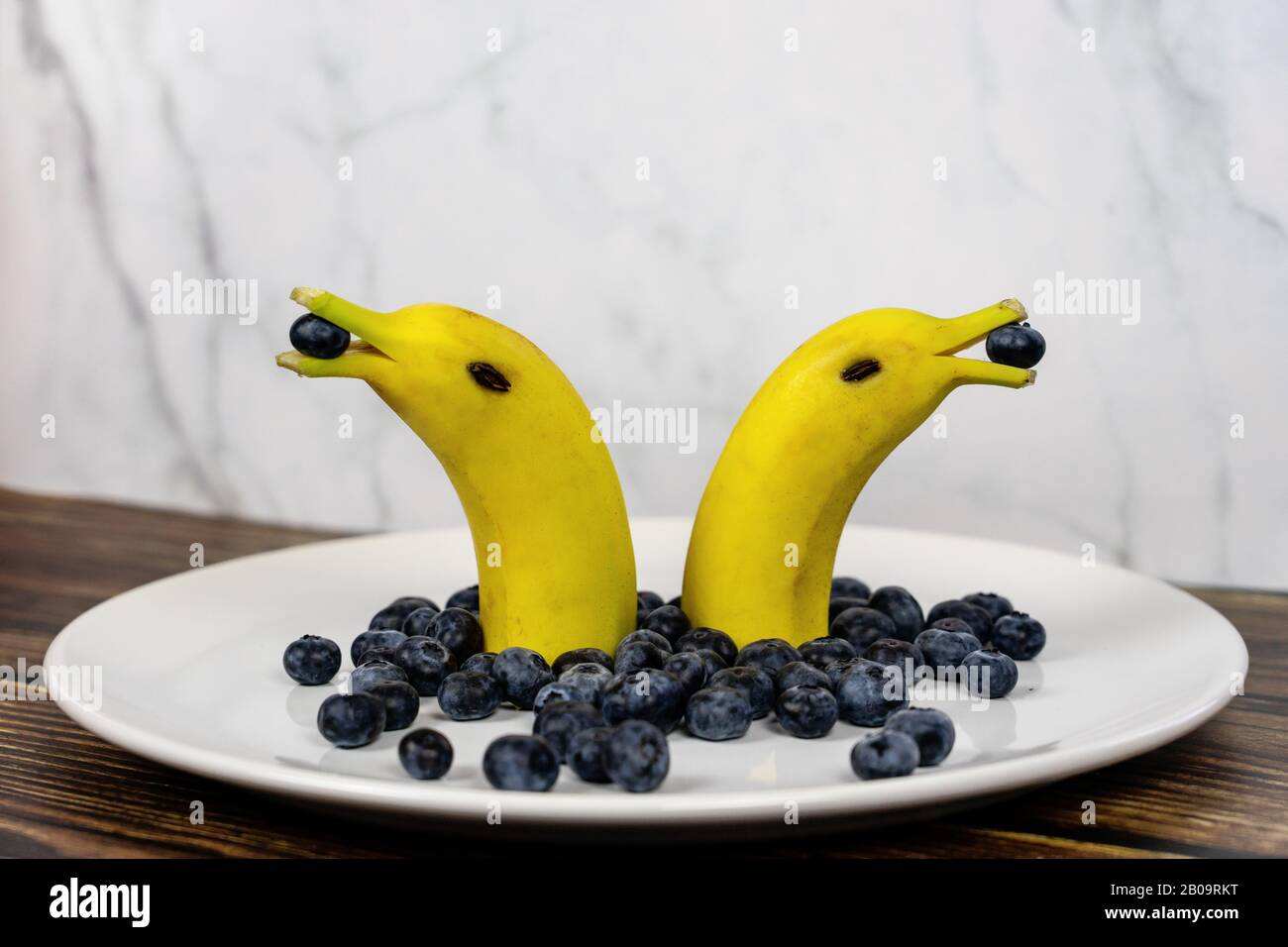 Lustige Obstfigur zwei Bananendelfine mit Blaubeeren auf einem weißen Teller. Stockfoto