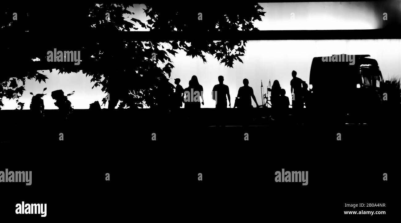 Silhouetten von Baumzweig und Jugendlichen in der Sommernacht, vor einem modernen Gebäude mit geparkten Motorrädern und einem Kleintransporter, in kontrastreicher Bla Stockfoto