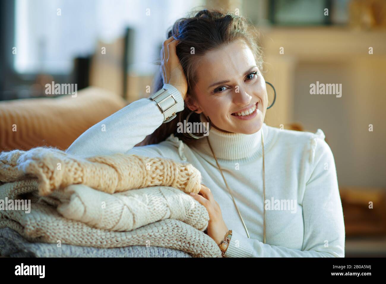 Porträt einer lächelnden, stilvollen Frau in weißem Pullover und Rock in der Nähe der Couch mit einem Haufen Pullover im modernen Wohnzimmer am sonnigen Wintertag. Stockfoto