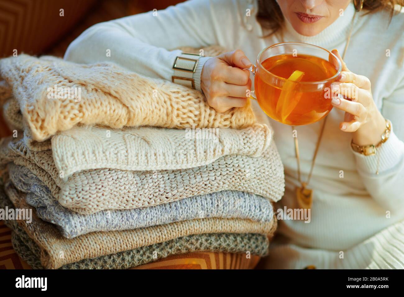Im modernen Wohnzimmer am sonnigen Wintertag können Sie sich im eleganten weißen Pullover und Rock mit Pullover, die im modernen Wohnzimmer eine Tasse Tee mit Zitrone trinken, ausziehen Stockfoto