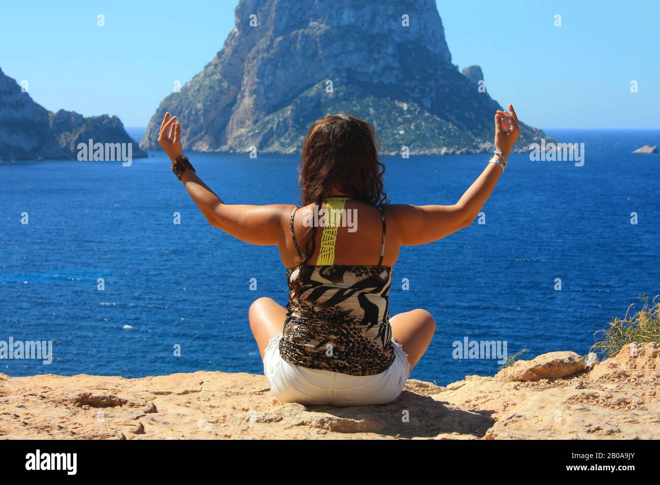 Das kaukasische Mädchen entspannt sich, indem es im Sommer in Cala Dhort buddhistische Meditationen auf einer Klippe vor der Insel Es Vedra in Spanien am Meer unternimmt Stockfoto