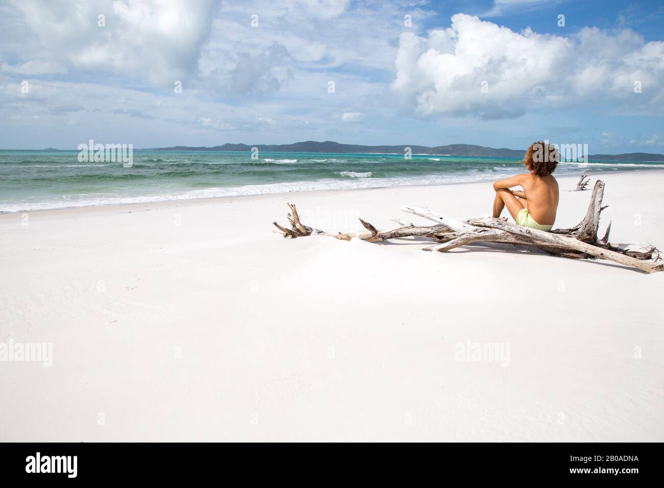 Mann mit lockigen Haaren, auf Holzschrammboden am weißen Sandstrand von Australien sitzend Stockfoto