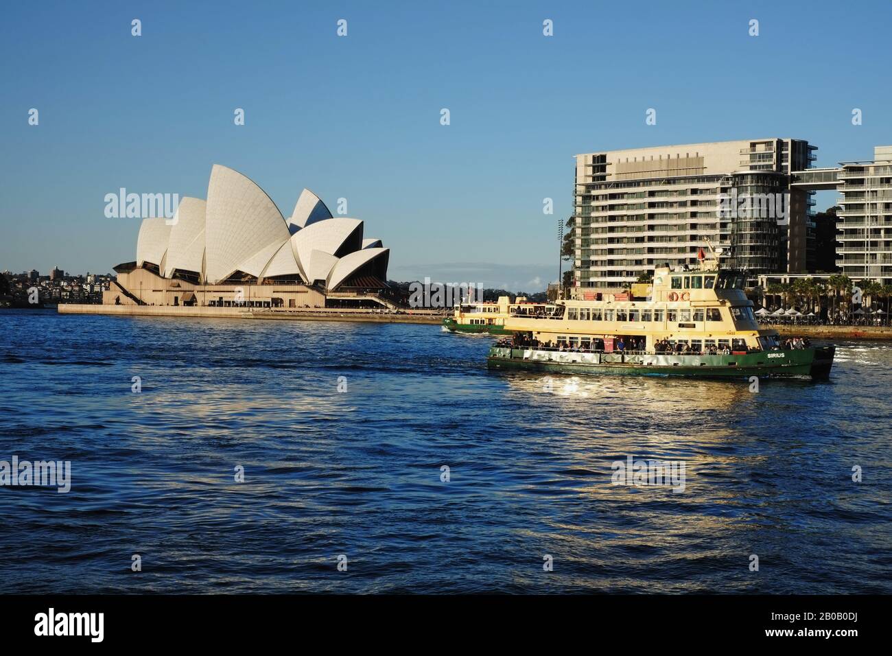 Sydney Opera House in sanftem Nachmittagslicht mit gelben und grünen Fähren, die sich unter blauem Himmel zum Circular Quay-Kai begeben Stockfoto