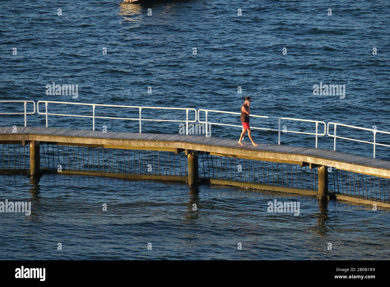 Sydney Summer - EIN Mann in Shorts und Singleausreden auf einem Handy, während er auf einem Boardwalk über blaues geripptes Wasser am Murray Rose Pool spazieren geht Stockfoto