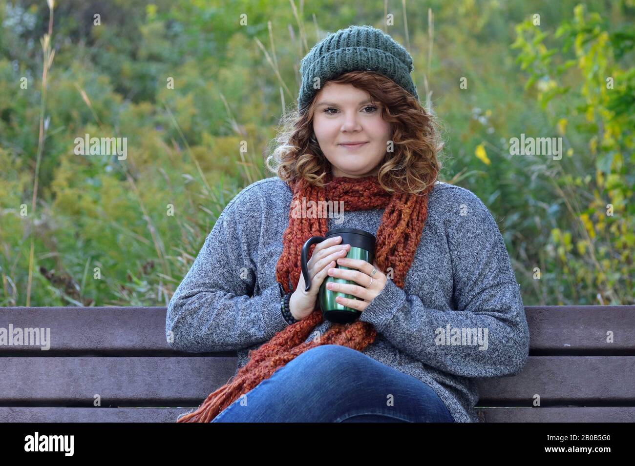 Eine junge Frau mit roten Haaren, die ein warmes Getränk genießt, während sie auf einer Parkbank sitzt. Stockfoto