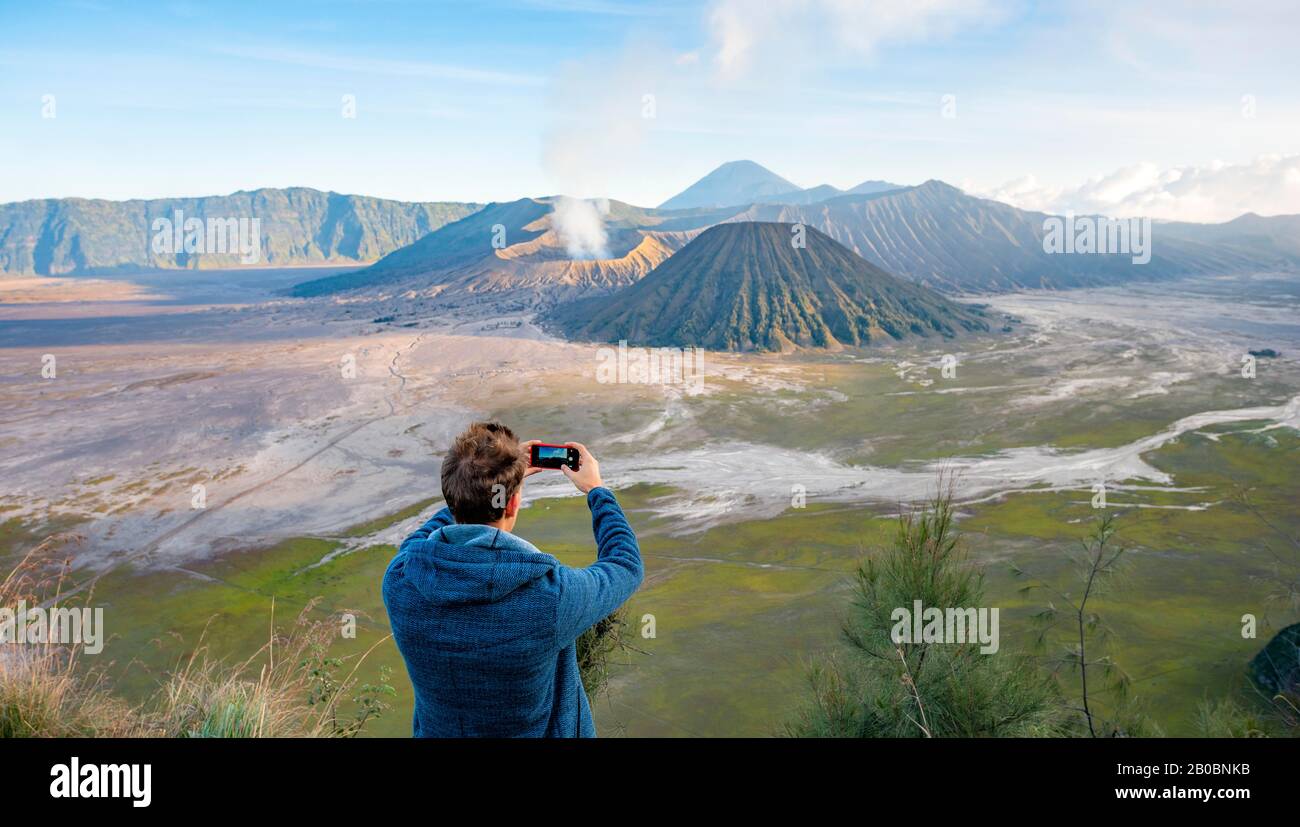Junger Mann fotografieren mit iPhone, Vulkanlandschaft, Blick in Tengger Caldera, rauchender Vulkan Gunung Bromo, vor Mt. Batok, im Rücken Mt. Stockfoto