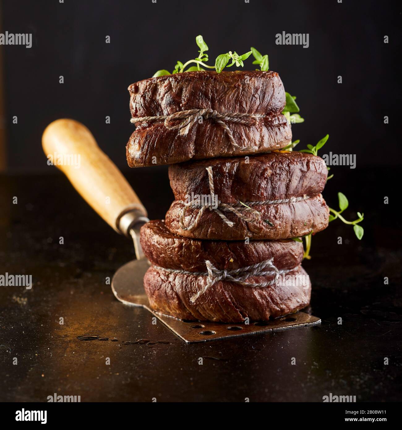 Feinschmecker dicke saftige gegrillte Steakmedaillons, die auf einem Metallspachtel zusammengestapelt und mit frischen Kräutern in quadratischer Form auf der Achäne garniert wurden Stockfoto