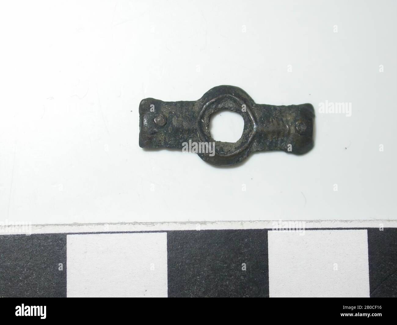 Balken, dunkel mit goldfarbenen Flecken (Kupferlegierung). Stange mit einem Loch (Dm 0,4 cm) in der Mitte. Der Rand um das Loch ist breiter als der Balken. Durch die Enden befindet sich ein Stift. Striche über die Breite des Strahls zwischen den Enden und der Bohrung. Der Rücken ist flach. 2-polige Montage. Stift ist nicht messbar., Bandhub, Metall, Kupferlegierung, L: 1,8 cm, W: 0,8 cm, D: 0,15 cm, Ende des 14. Jahrhunderts 1350-1400, Niederlande, Zeeland, Holly, Verdronken Land Stockfoto