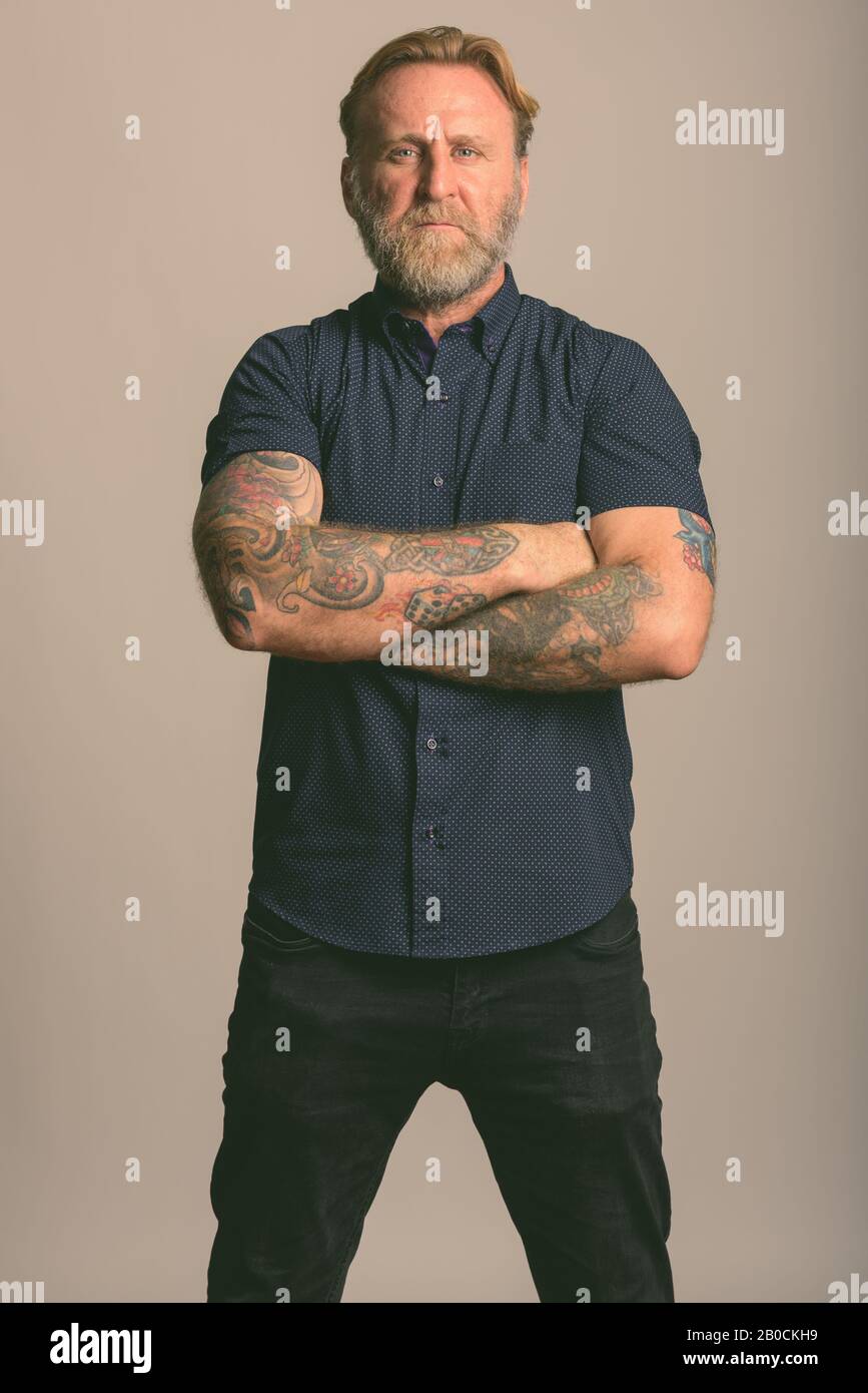 Studio shot reifer bärtiger Mann mit hand Tattoos stehend mit Waffen in grauer Hintergrund gekreuzt Stockfoto
