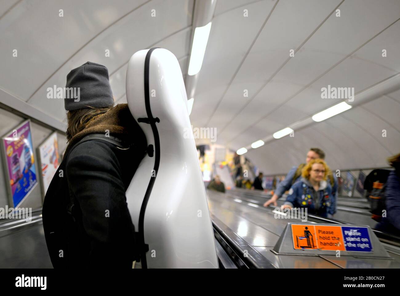 London, England, Großbritannien. Mann, der ein Cello in einer U-Bahn-Station eine Rolltreppe hinunterträgt Stockfoto