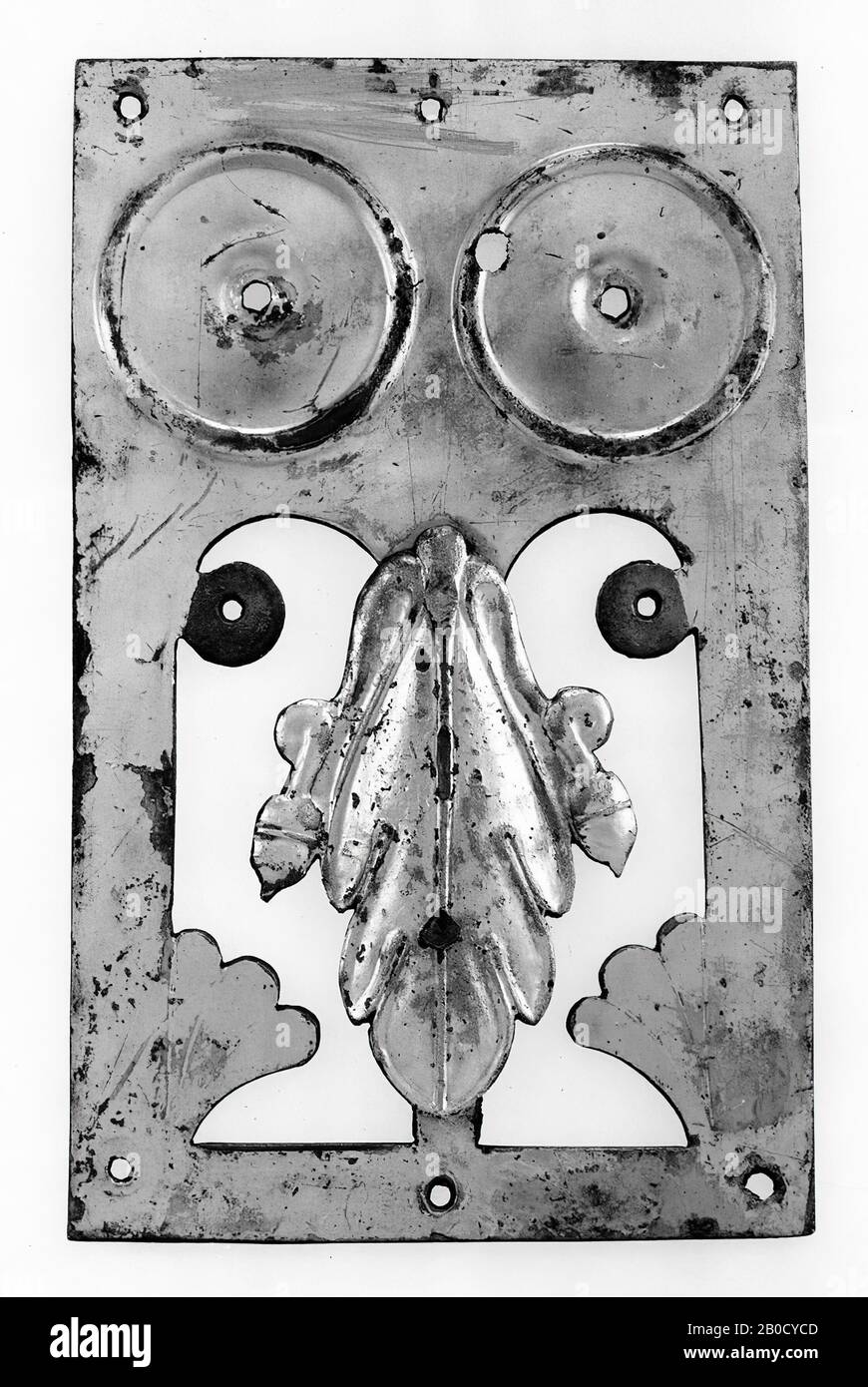 Rechteckige versilberte Bronzeplatte. Oben befinden sich zwei runde Verzierungen (mit einem konkaven Mittelfeld, das ein Lochfeld umschließt), um die ein Rand, zuerst konvex als konkave, geschlossen von einem dicken Grat) herausgetrieben wird. Darunter befindet sich ein großes dekoratives Motiv. Zwei längliche Bogenfenster sind nebeneinander ausgeschnitten. An den Außenseiten der Fenster verbleiben ein Kreis (mit einem Loch in der Mitte) und unten ein fächerförmiges Muster in Bronze. Auf dem dünnen, dazwischen liegenden Stil, der die beiden Fenster voneinander trennt, ist eine Zierde verlötet: Ein konvexes Eichenblatt mit einer Akorn auf beiden Seiten. Hinten sind sie kurz Stockfoto