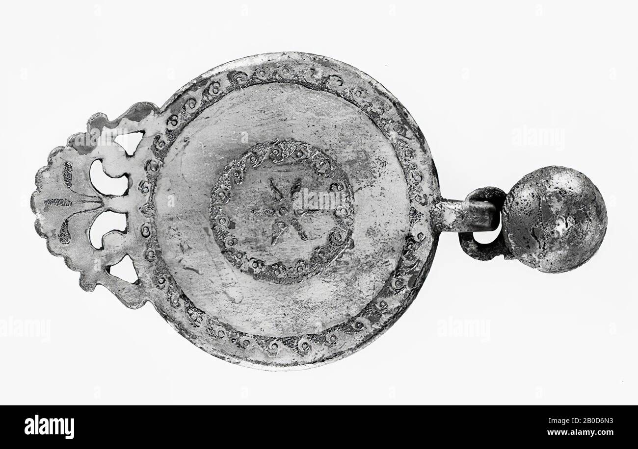 Flacher runder Anhänger aus versilberter Bronze, der oben einen Haken hat und unten ein sichelförmiges, gezähntes Ziermotiv. Der Haken ist an einem ca. Rechteckigen Stück Bronze befestigt, in dem ein Auge sitzt. Der Haken wurde entfernt. In der Mitte des Bronze-Stücks befindet sich ein Loch, in dem mittels eines Stifts eine konvexe Scheibe (eine Art Knopf) befestigt ist, die mit einem Ring gehalten wird. Anhänger der Dekoration: In der Mitte stehen 6 Blätter, die von einem Mittelpunkt enden. Um ihn herum befindet sich ein Ring mit Wellenmotiv, genau wie der Rand des Pendants. Im gezackten Schmuckmotiv ein Dreieck Stockfoto