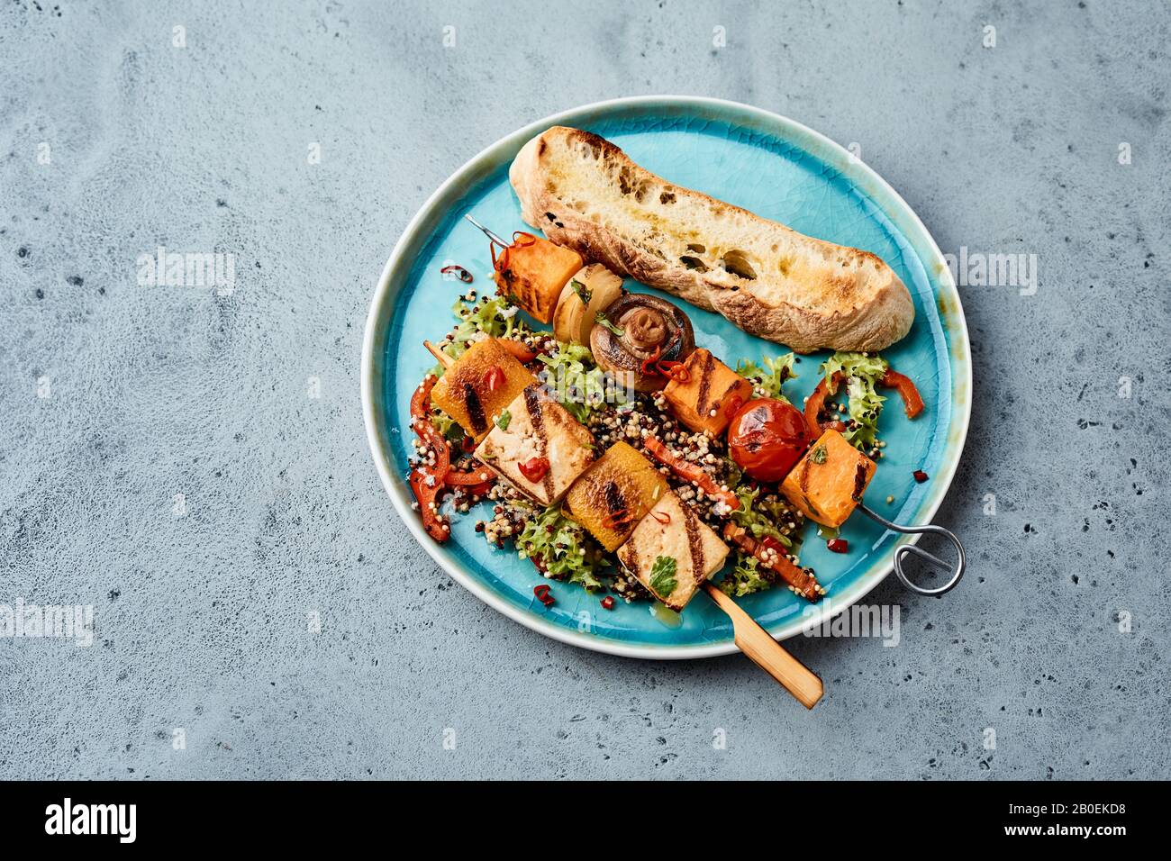 Leckere, gegrillte Gemüsekebabs mit Tofu, Kürbis oder Butternuß, Tomaten und Pilzen auf einem Bett aus Salatgrüns, das auf einem blauen Teller im Overhead serviert wird Stockfoto