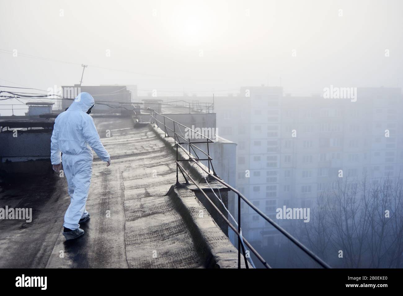 Rückansicht des Mannes, der weißen Overall und Gasmaske trägt und auf dem Dach eines städtischen Gebäudes spaziert. Vor dem Hintergrund der aufgehenden Sonne bei nebeligem Wetter, Konzept des Umweltproblems Stockfoto