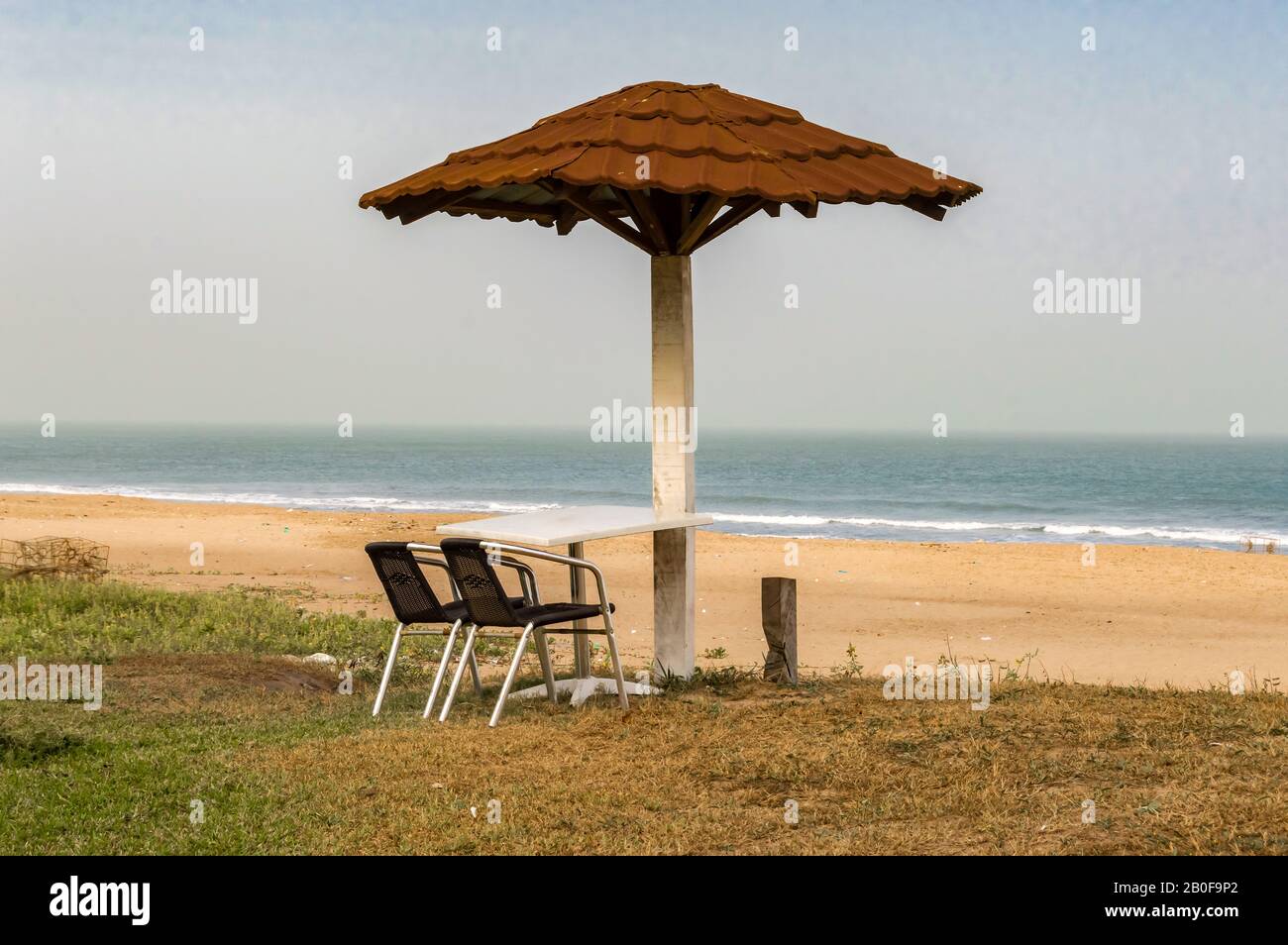 Zwei Stühle und ein Tisch liegen zum Meer, knapp unter einem roten Fliesenschirm an einem Strand in Gambia Stockfoto