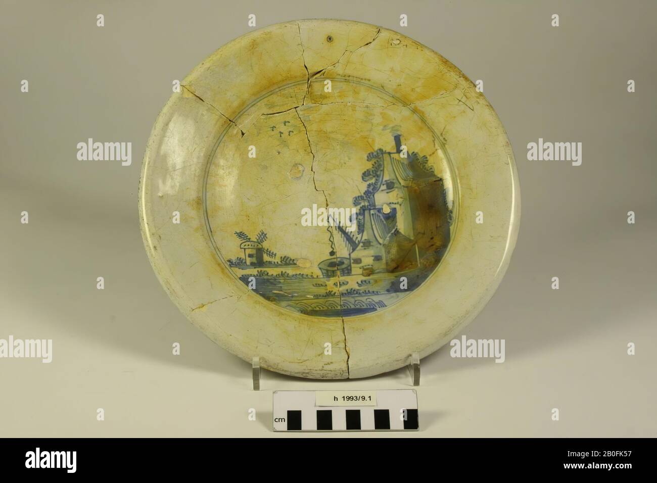 Platte mit blauem Faience-Gemälde. Nur der Spiegel ist mit blauem Kobalt bemalt. Es geht um die Darstellung eines Hauses mit glockenförmigem façade mit rauchenden Schornsteinen und umsäumt von Bäumen, davor ein bauchiges Haus gleichen Typs. Neben den Häusern gibt es einen Brunnen, eine kleine Mühle am Wasser und drei Vögel in der Luft. Dating: Zweite Hälfte des 17. Jahrhunderts. Alte Klebepunkte, gelbe Flecken auf der Oberfläche, knistern auf der Oberfläche, müssen auf Spiegel, Platte, Steingut, Faience, rotes Backen, h: 5,4 cm, Durchm: 30 cm, nt 1650-1700, Niederlande, Südholland, Alphen aan den Rijn stehen Stockfoto