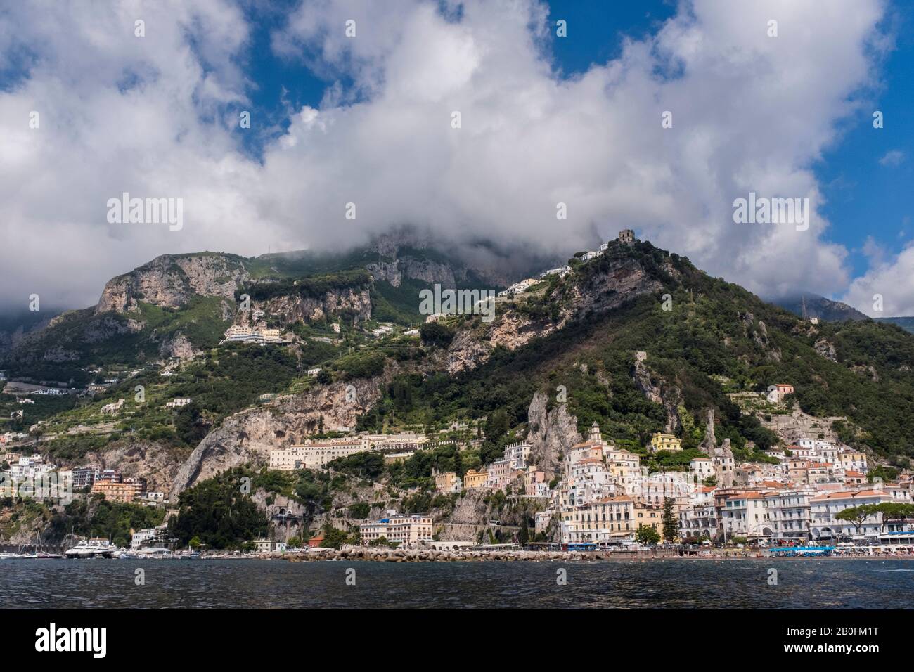 Das bunte Dorf Amalfi, vom Meer aus gesehen, mit dramatischen Bergen und sommerlichem Himmel im Hintergrund, an der Küste Italiens Stockfoto