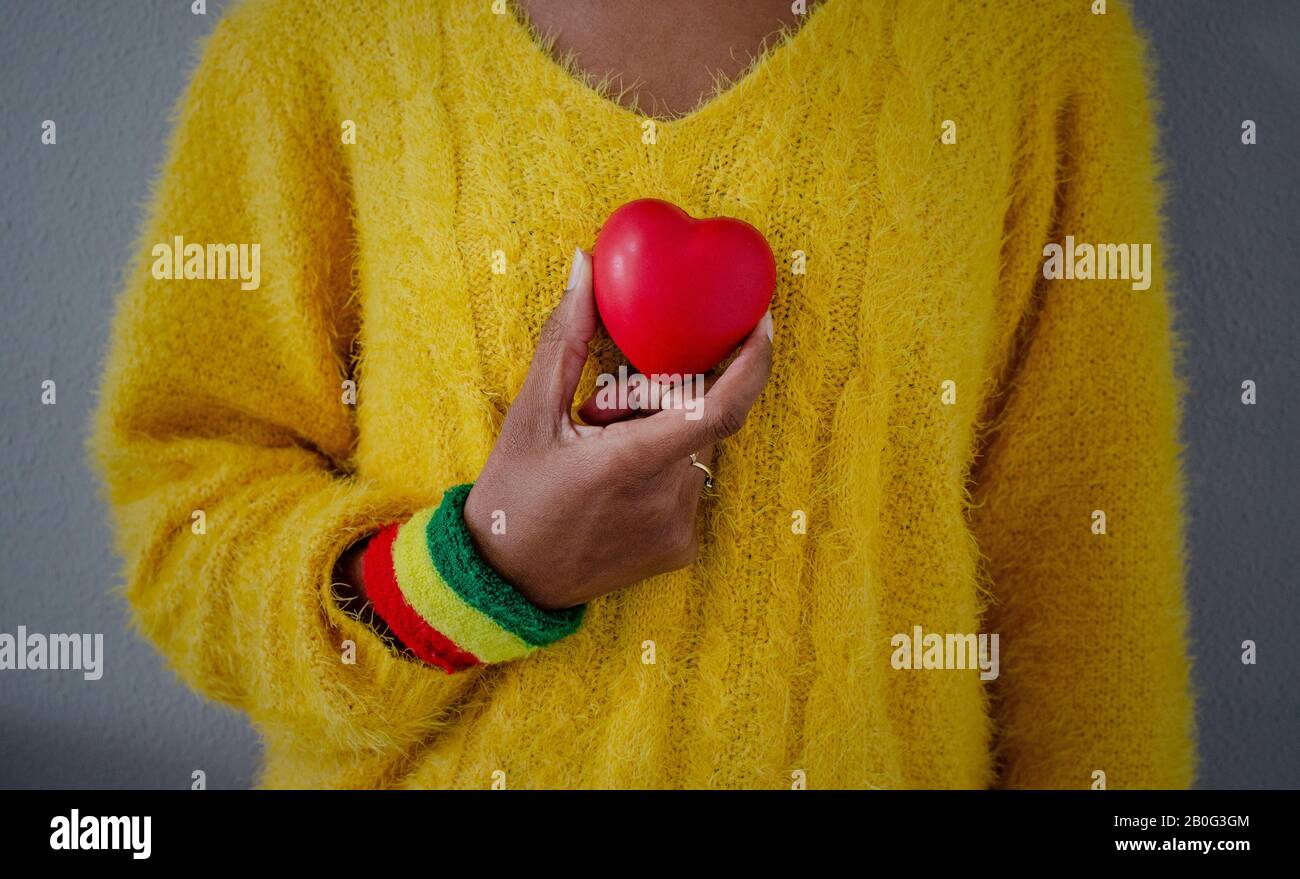 Hand einer schwarzen Frau mit einem roten, gelben und grünen Armband, das ein Herz gegen die Brust hält. Liebe, Gesundheitskonzept. Stockfoto