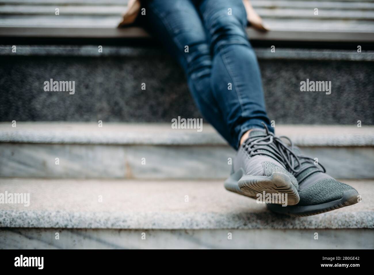 Nahaufnahme der weiblichen Beine mit dunkelblauen Jeans und Turnschuhen. Junge Frau, die auf Steinstufen im Stadtpark sitzt. Stockfoto