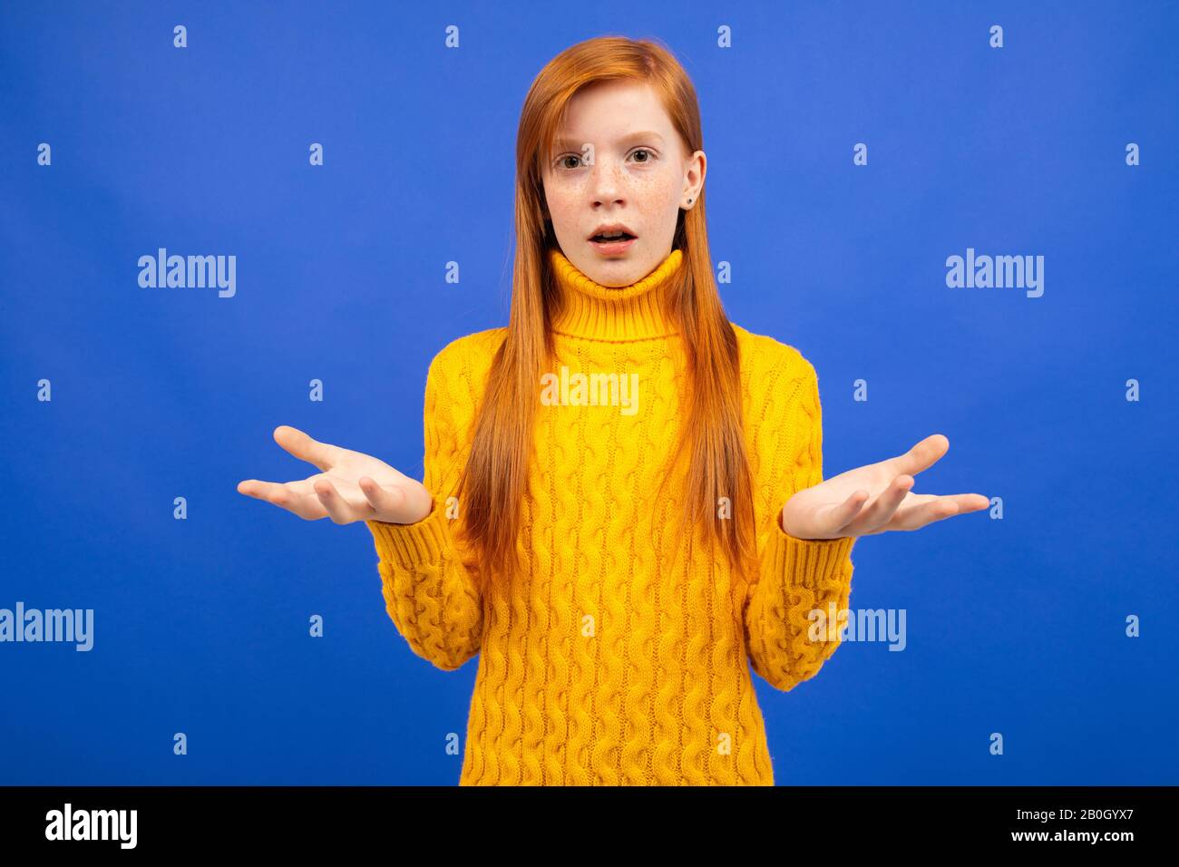 Charmantes kaukasisches rothaariges Mädchen in einem gelben Pullover, das ihre Arme schwenkt und vor einem blauen Studiohintergrund wieder auftaucht Stockfoto