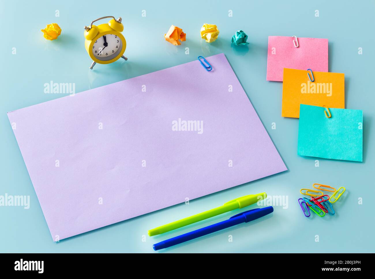 Gelber Wecker auf einer violetten Karte mit der Aufschrift "geschäftig Tag" farbige Büroklammern, Karten, Stifte, zerknitterte Papierkugeln. Platz für Text Stockfoto
