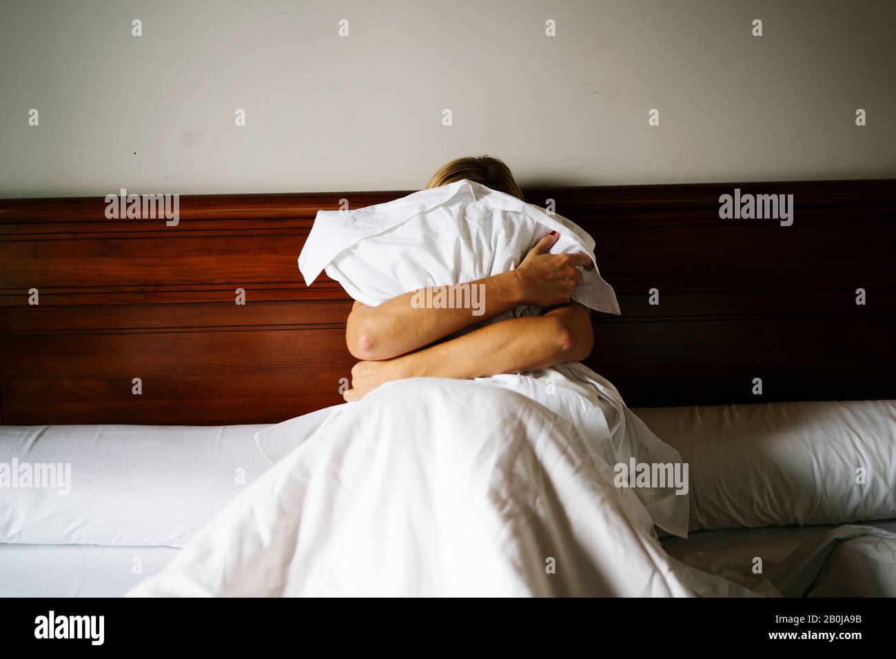 Ein blondes Mädchen, das auf dem Bett sitzt, bedeckt sich mit dem Kissen Stockfoto