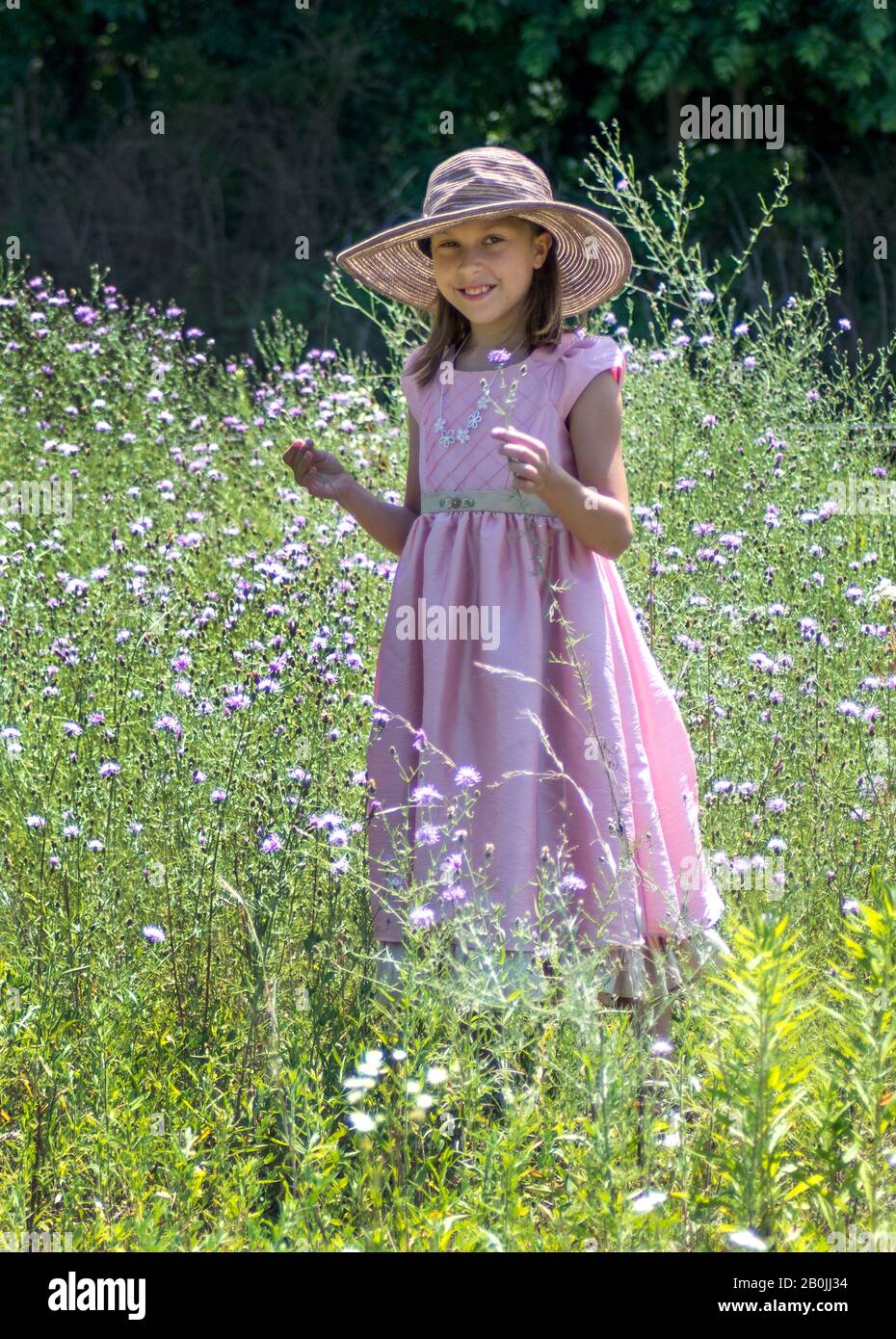 Lächelndes 10-jähriges Mädchen in einem hübschen, rosafarbenen Kleid und einem Floppyhut hält Blumen, während sie in einem Feld von Blumen steht Stockfoto