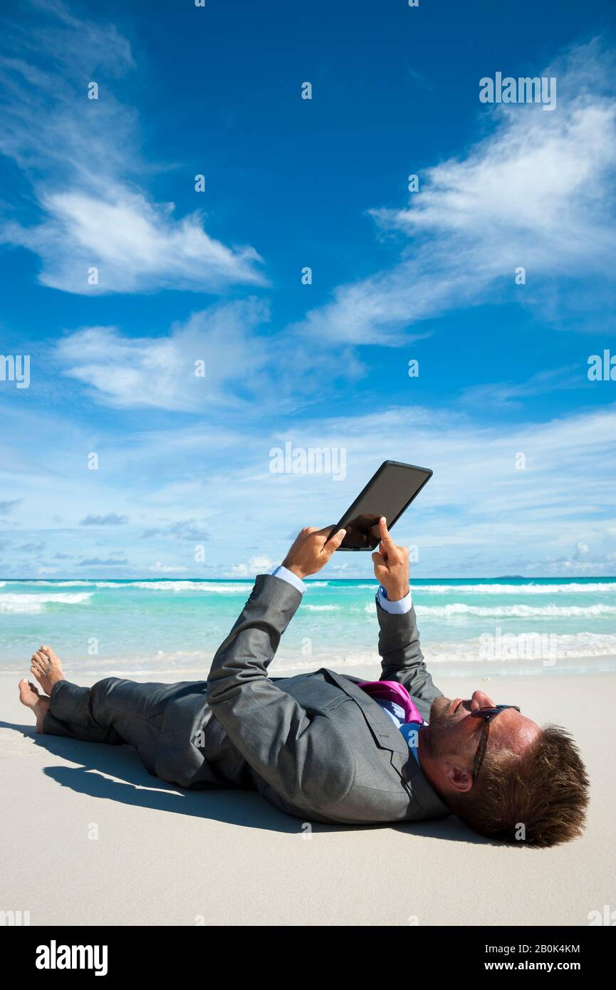 Entspannter Geschäftsmann, der mit seinem Tablet-Computer am Ufer eines tropischen Strandes in einem nassen Anzug liegt Stockfoto