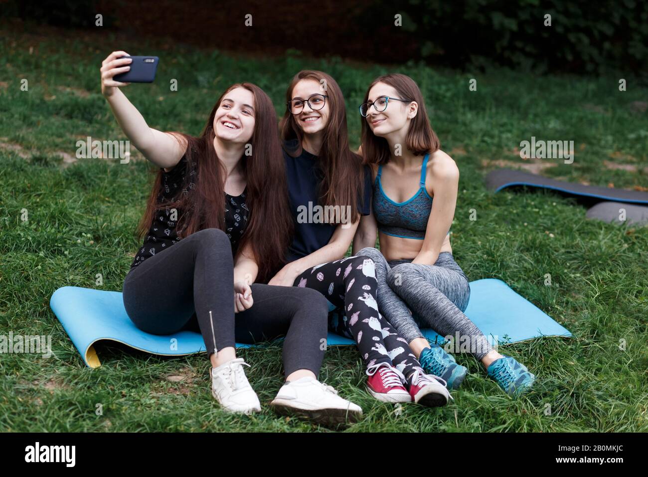 Drei Mädchen sitzen auf Yogamatte und machen Selfies im grünen Park Stockfoto