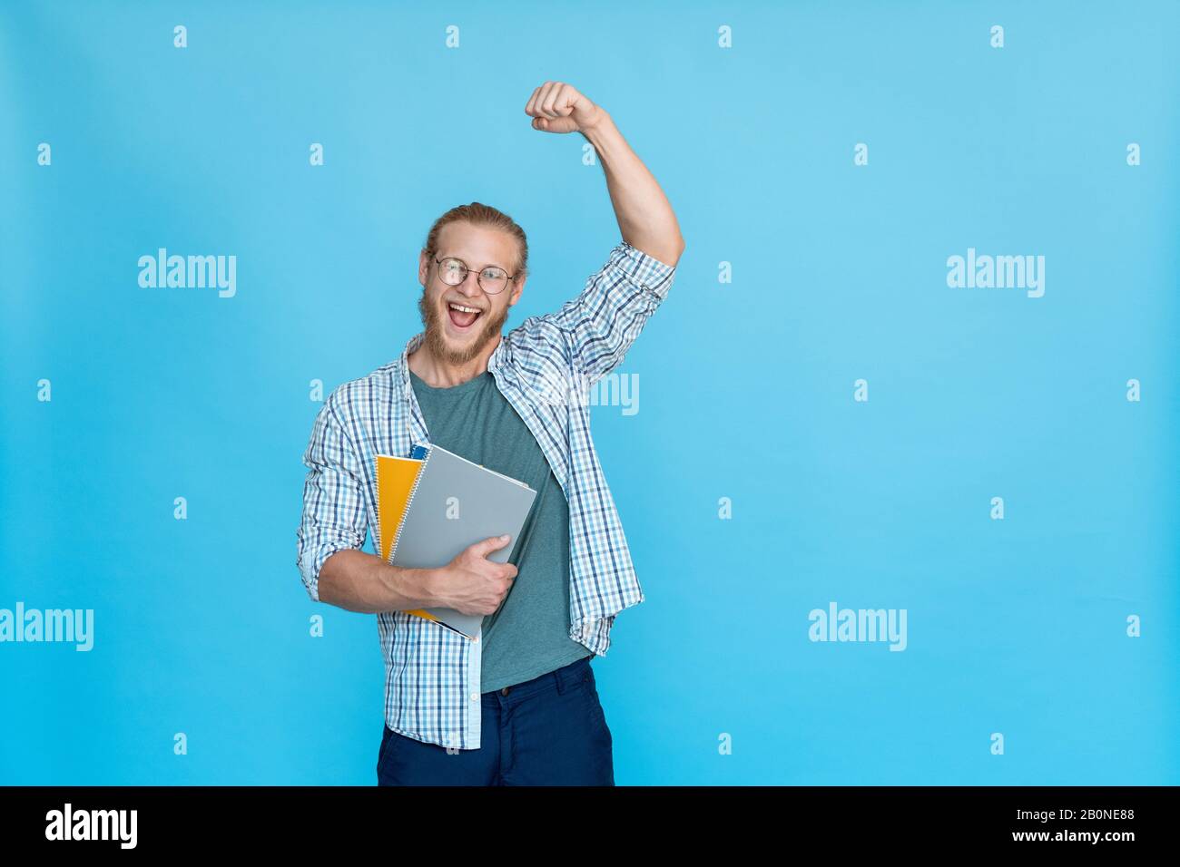 Bärtiger, glücklicher, aufgeregter Student in Brille hält eine Gewinn-Geste für das Copybook Stockfoto