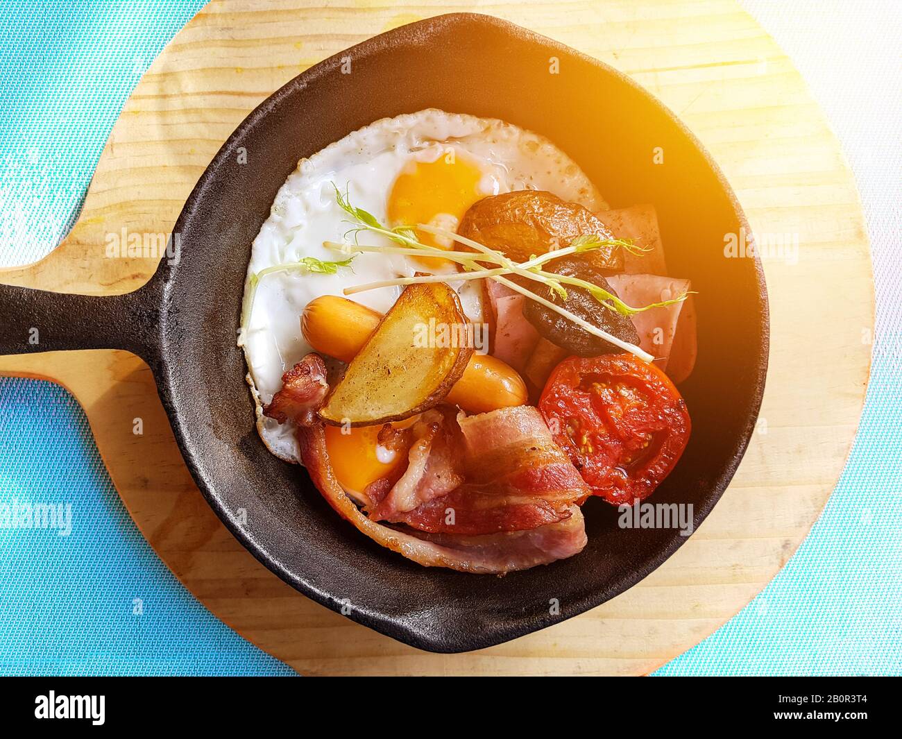 Frühstück in der Pfanne mit Spiegelei, Würstchen, Speck, Tomaten, Kartoffeln und Gemüse auf Holzblock Hintergrund auf dem Tisch in der Küche. Stockfoto