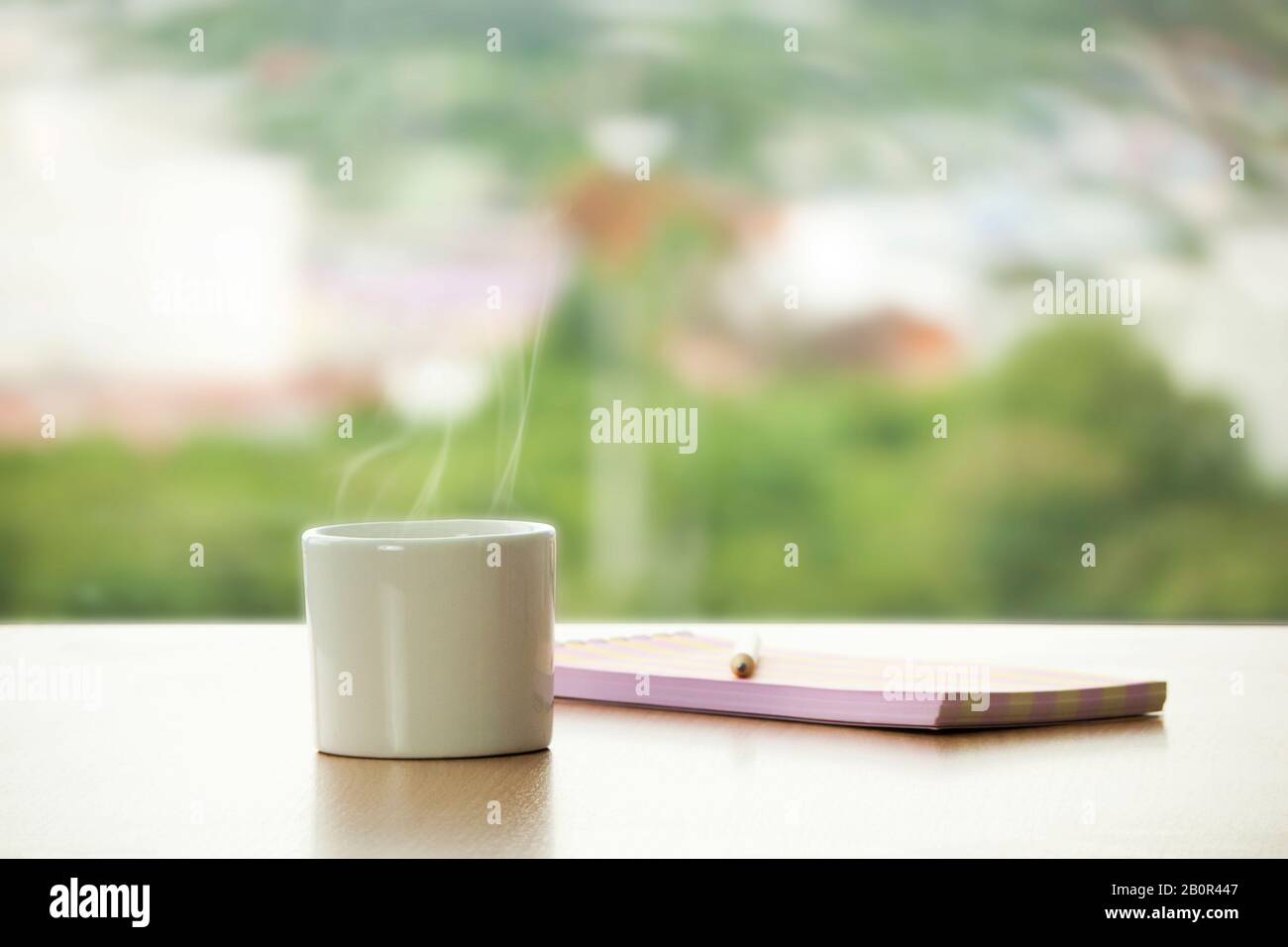 Tasse heißen Kaffee mit Bleistift und Notizbuch auf dem Tisch im Café. Café-Fensteransicht mit grünem Baum für frisches Gefühl als Hintergrund. Kreativ und Idee Stockfoto