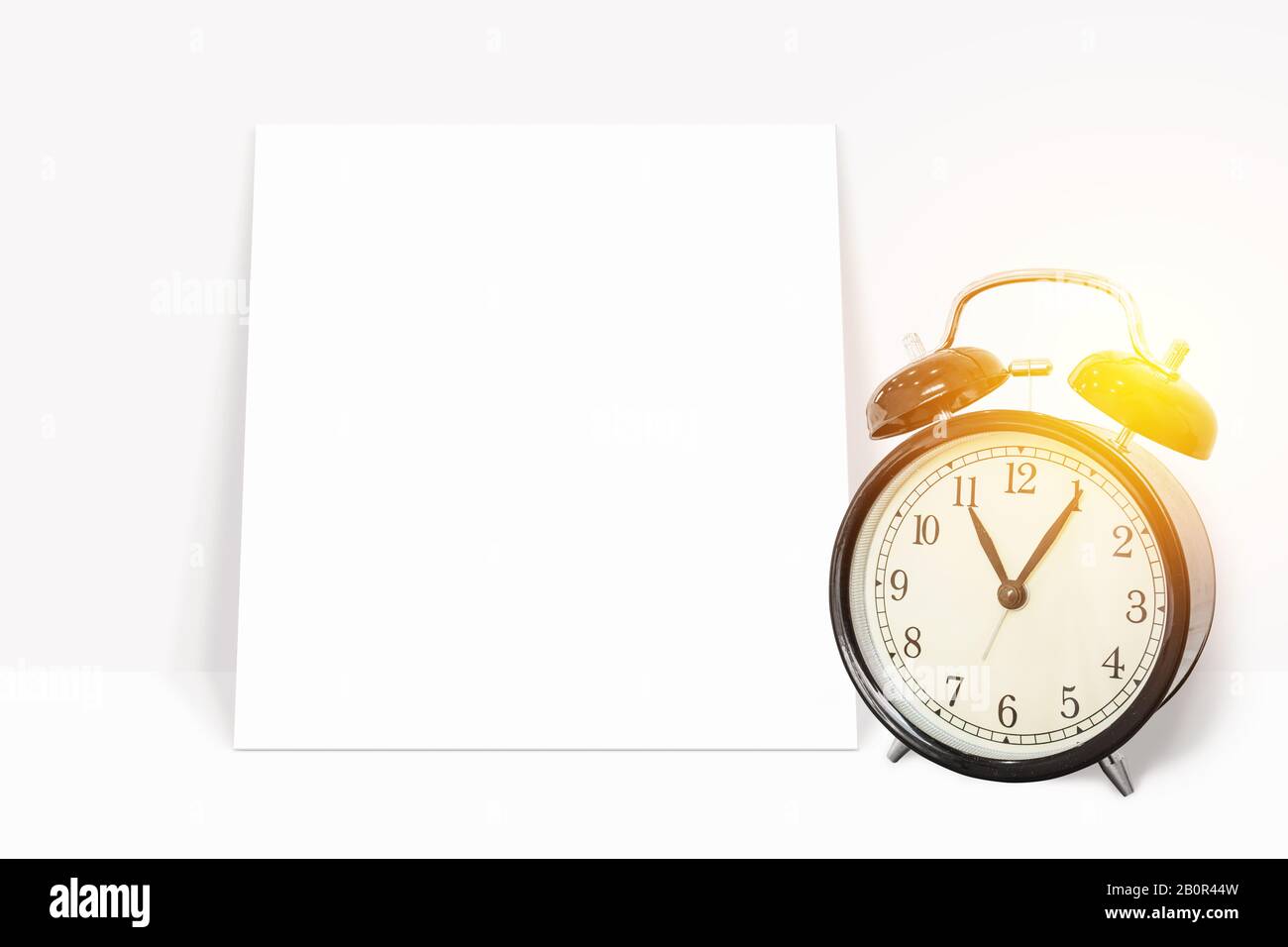 Zeitmanagement-Konzept: Analoge schwarze Uhr im Vintage-Format mit leerem White Paper an die weiße Wand gelehnt. Leeres White Paper mit Kopierplatz für den Stockfoto