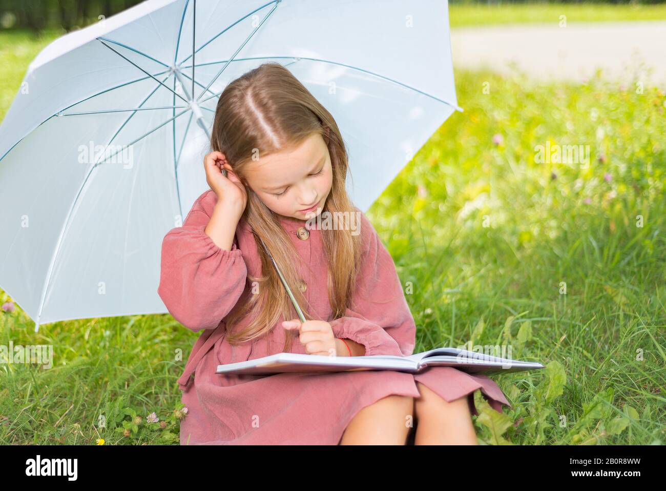 Kind liest ein Buch, kleines Mädchen unter einem Regenschirm sitzt an einem sonnigen Tag auf grünem Gras Stockfoto