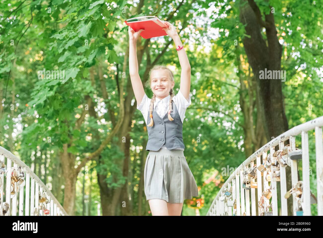 Student Schülerin mit Zöpfen in Uniform mit Bücher in die Hände über dem Kopf läuft glücklich über die Brücke im Sommer Park Stockfoto