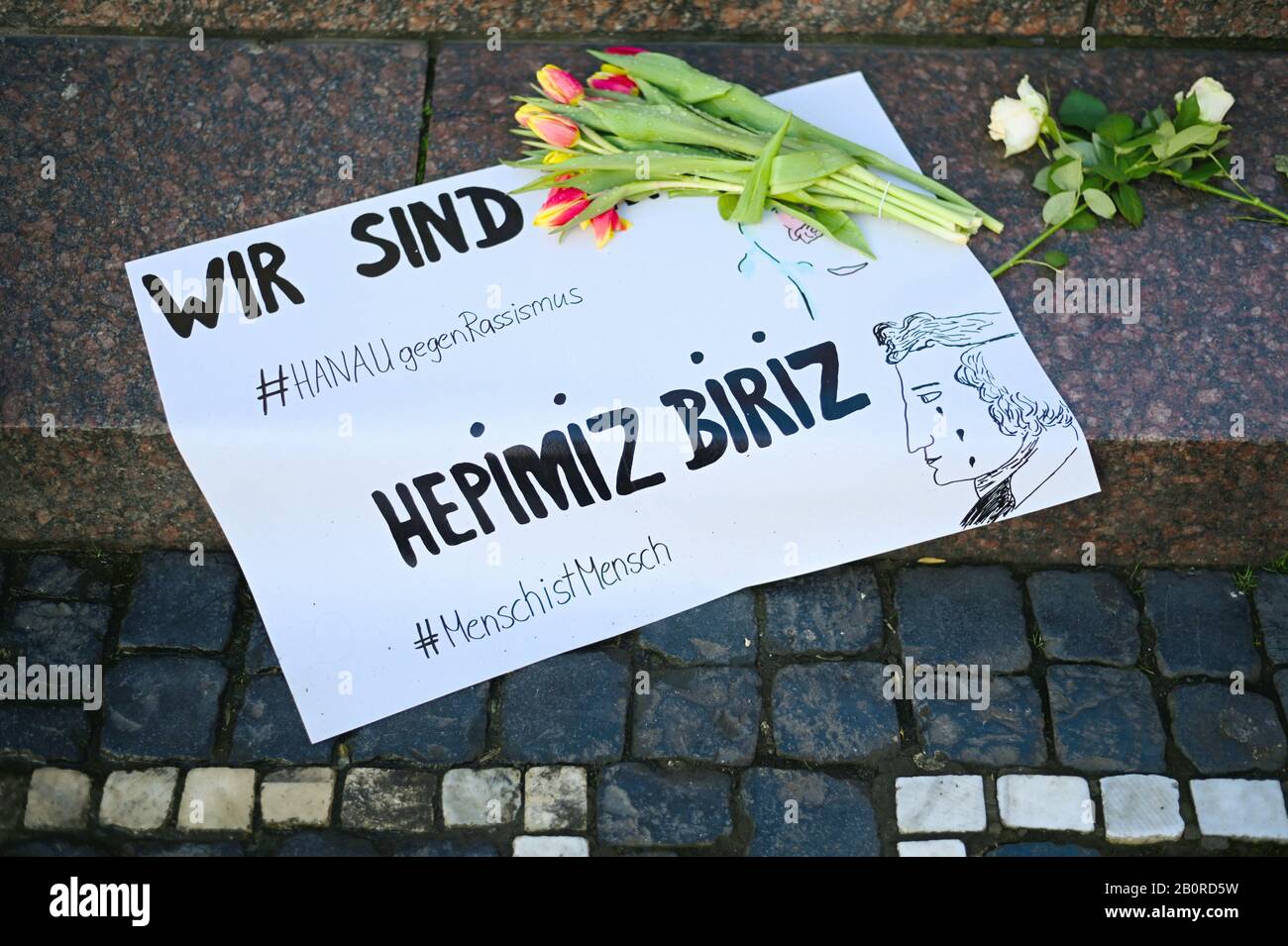 Hanau, Deutschland. Februar 2020. Blumen wurden auf dem Marktplatz neben einem Zettel mit den Aufschriften "Wir sind #HANAUagainstRacism", "HEPIMIZ BIRIZ" (türkisch für "Wir sind alle eins") und #MenschistMensch platziert. Bei einem angeblich rassistischen Angriff erschoss ein 43-jähriger Deutscher im hessischen Hanau mehrere Menschen und sich selbst. Credit: Nicolas Armer / dpa / Alamy Live News Stockfoto