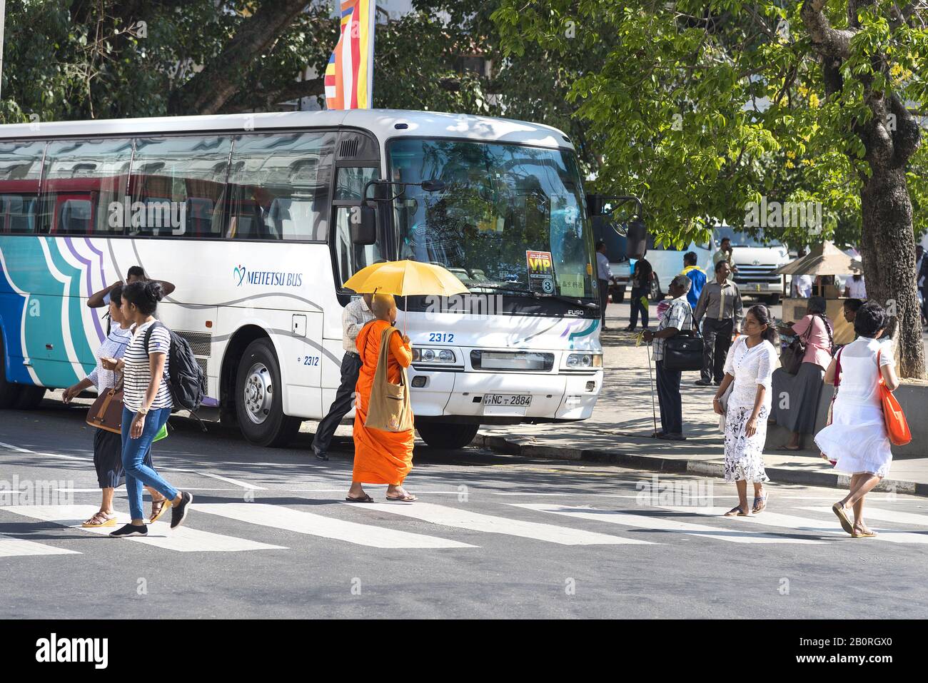 Kandy, Sri Lanka: 19.03.2019: Stadtzentrum mit Touristenreisebus und buddhistischem Mönch, der die von gelbem Regenschirm beschattete Straße überquert. Stockfoto