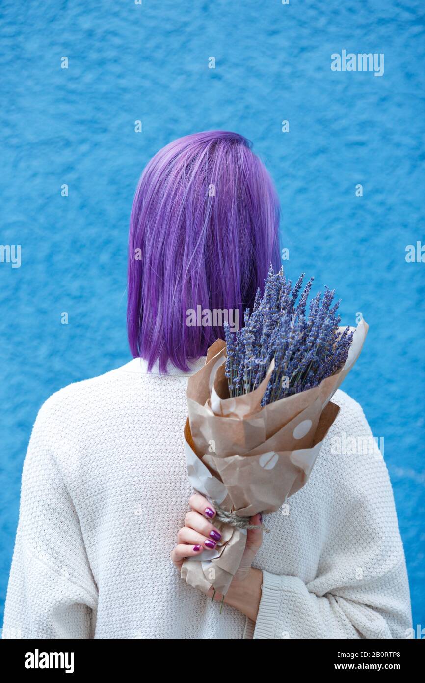 Junge Dame mit violettem Haar, weiße Bluse, stehend, mit Lavendelbouquet in Papierverpackung in der rechten Hand vor der blauen Wandrückengrou zurückdrehen Stockfoto