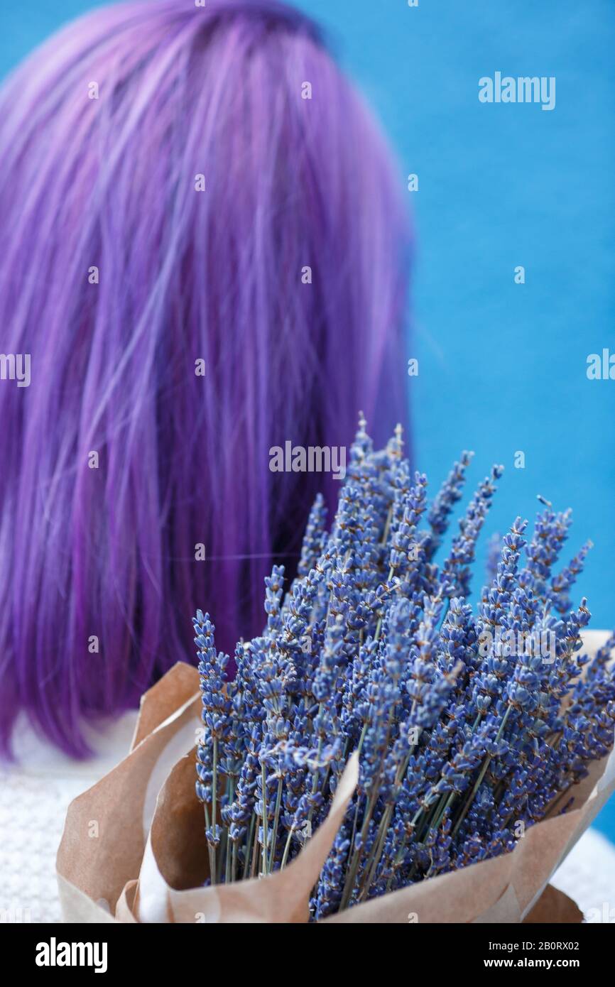 Hinterkopf einer Frau mit violettem Haar und Lavendelstrauß in Papierverpackung vor blauem Wandhintergrund Stockfoto