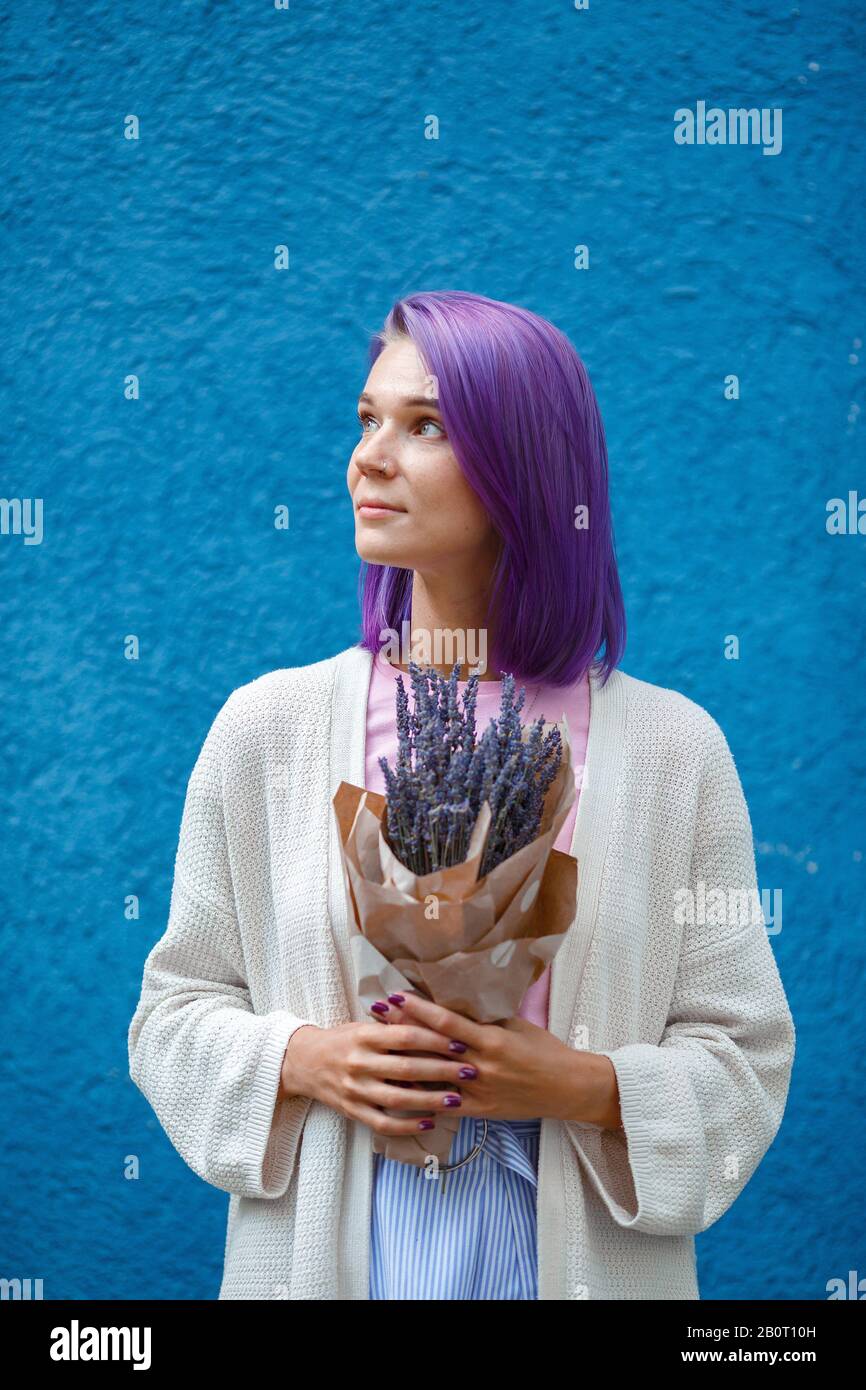 Portrait eines Mädchens mit violettem, farbenfrohem Haar, in weißer Strickjacke und blauem Rock, das einen Lavendelstrauß in Papierhüllen in ihren Händen hält. Mädchen steht Stockfoto