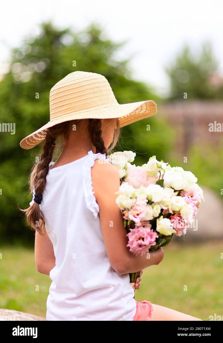 Mädchen mit einem Blumenstrauß aus weißen und rosafarbenen Ponys. Sonniger Sommertag. Mädchen in einem Hut mit breitem Rand. Rückansicht. Mädchen mit zwei Zitzen. Stockfoto
