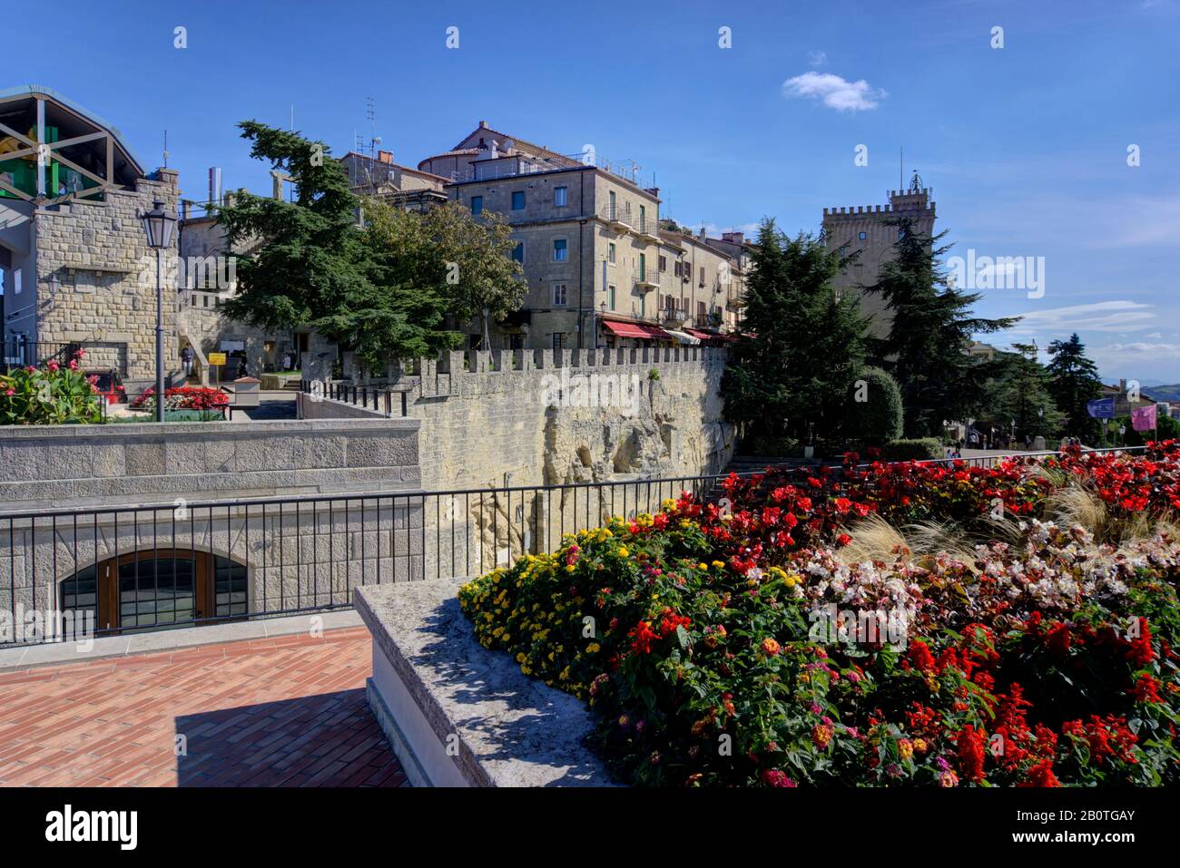 San Marino, San Marino - 19. Oktober 2019: Landschaftlich schön über die Aussichtsplattform in Richtung alter, steinerner Bauwerke, die bunte Blumen und Touristen zeigen Stockfoto